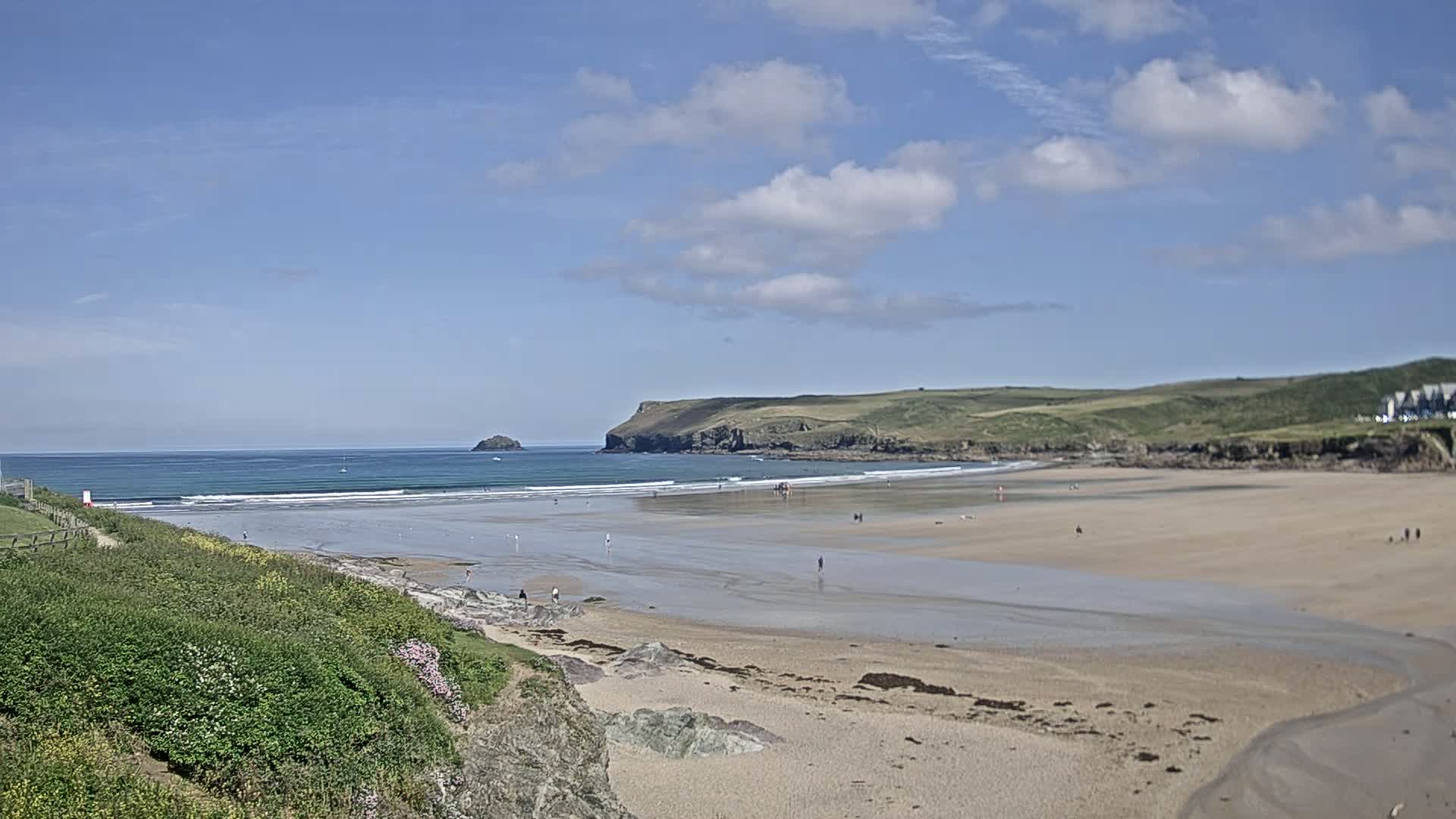 A sandy beach stretches out to a calm ocean under a partly cloudy blue sky, with a few people scattered on the sand and small waves rolling in.