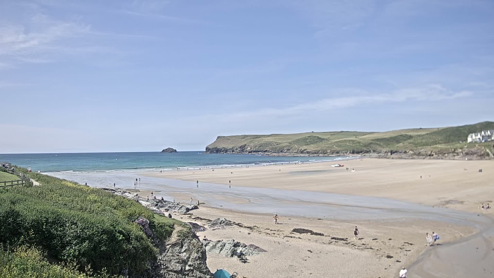 A sandy beach stretches out to a calm ocean under a clear blue sky, with a few people scattered across the sand and in the water.