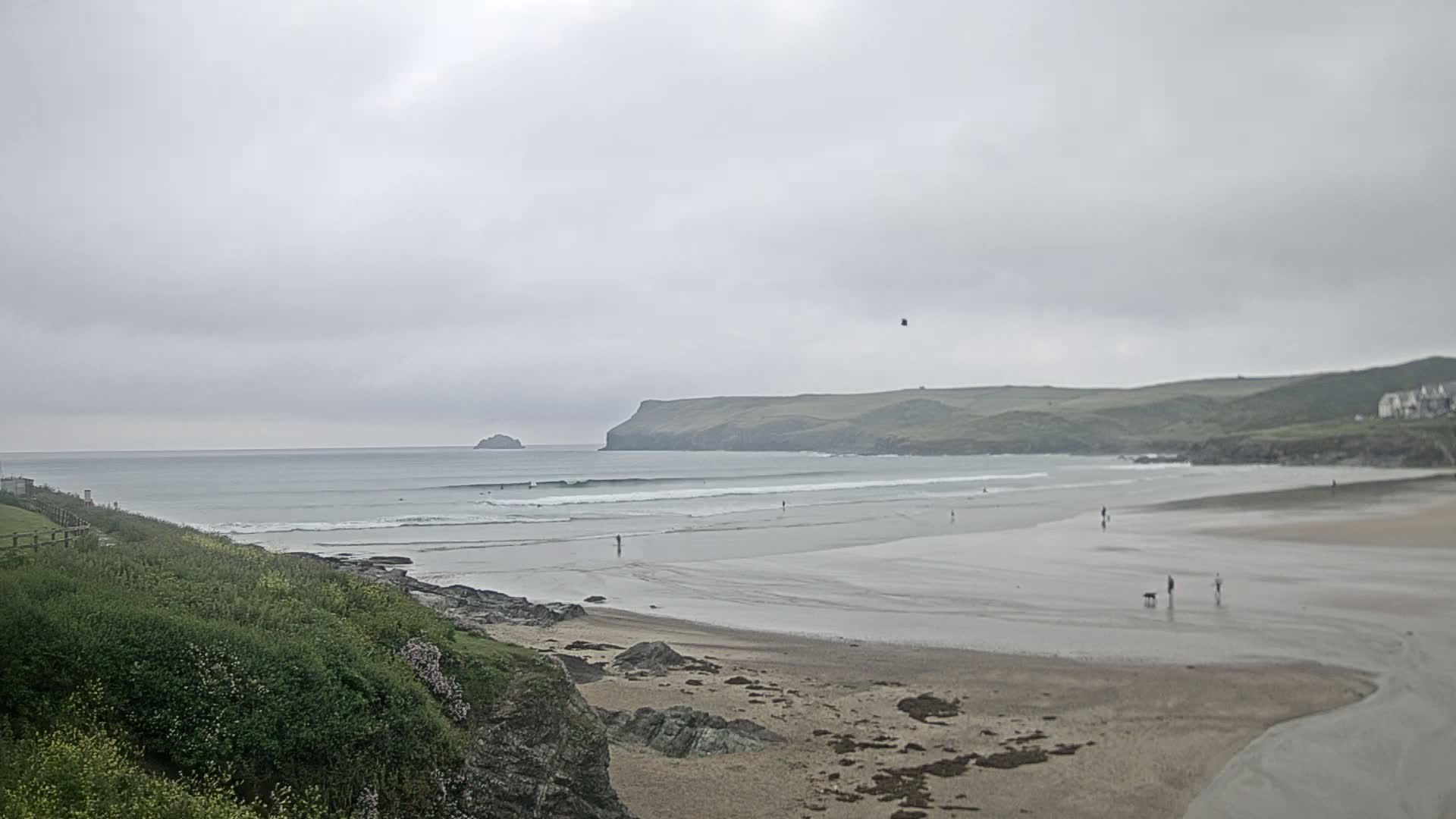 A wide sandy beach stretches out to a calm ocean under an overcast sky, with a few people walking on the sand and some surfers in the water.