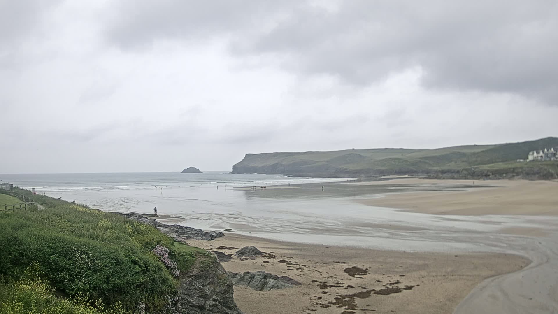 A sandy beach with a calm ocean under an overcast sky is seen from a grassy cliff overlooking the scene.