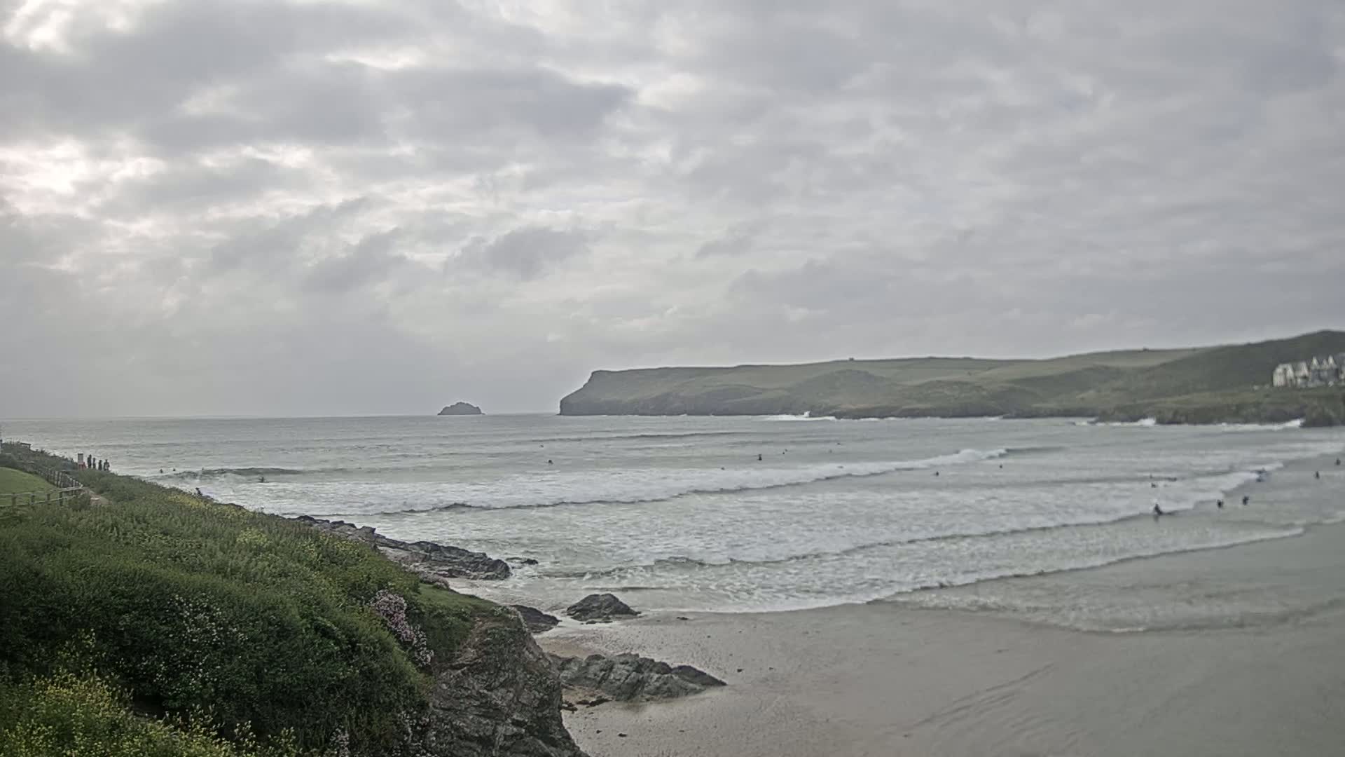 A beach with several surfers in the ocean under a cloudy sky is visible from a clifftop covered in green vegetation.