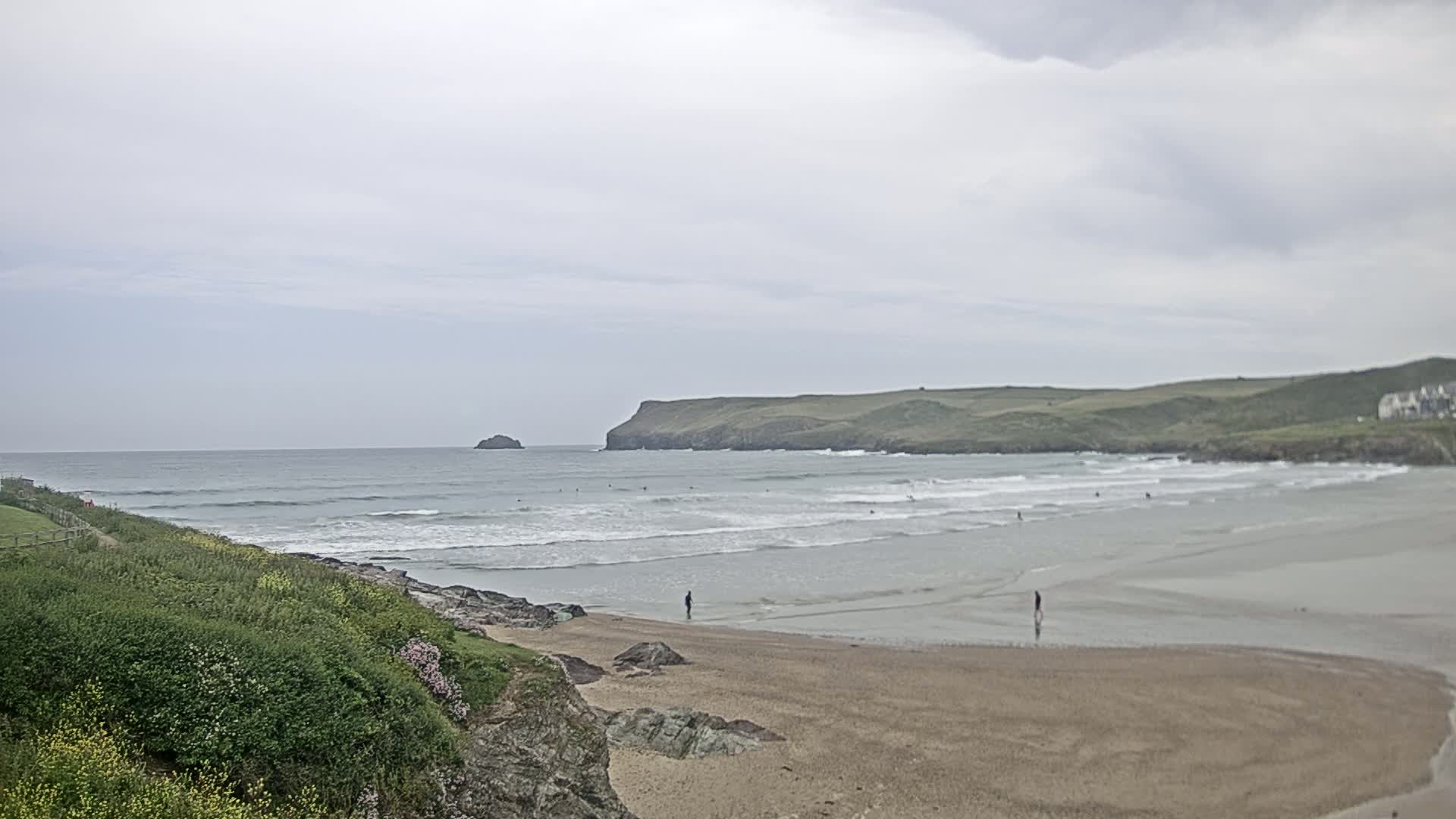 A sandy beach with gentle waves under an overcast sky, a few people are seen on the beach and in the water, with green hills and a small island in the distance.
