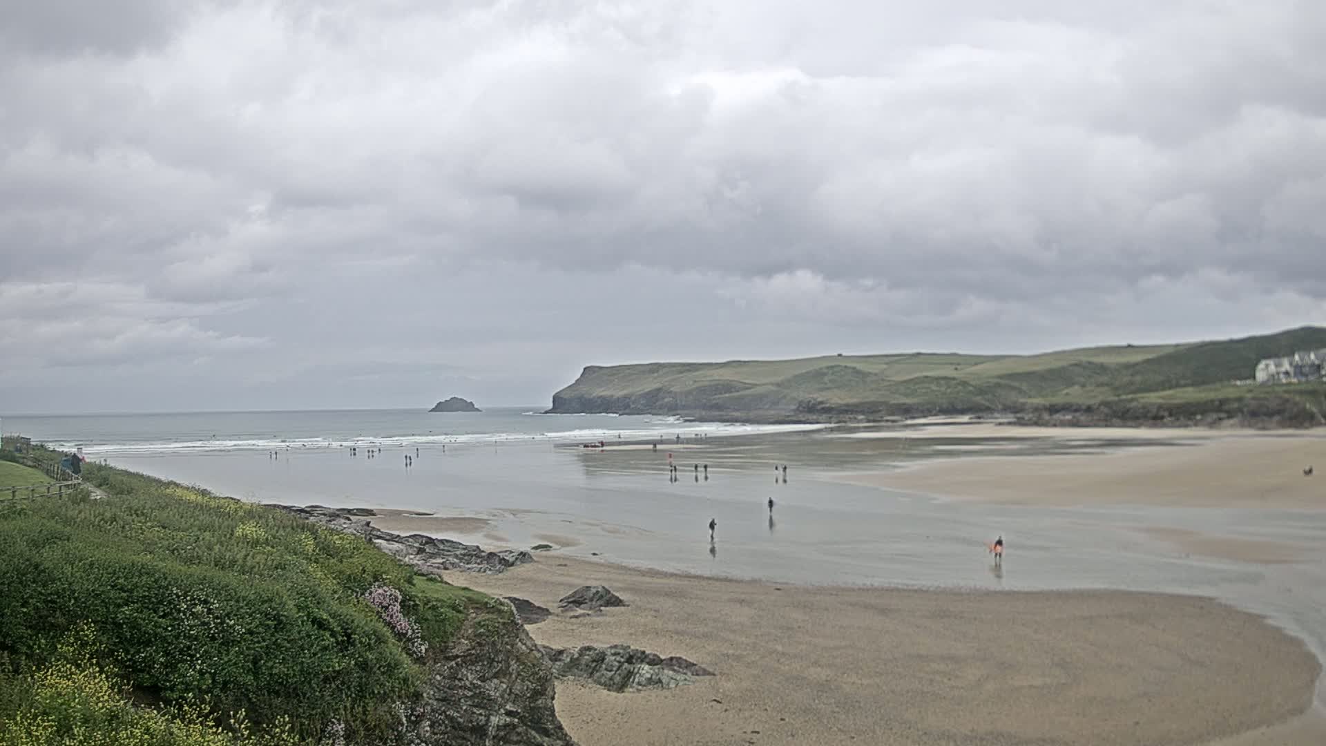 A wide sandy beach stretches out under a cloudy sky, with people sparsely scattered across the sand and in the surf, and a small island visible in the distance.