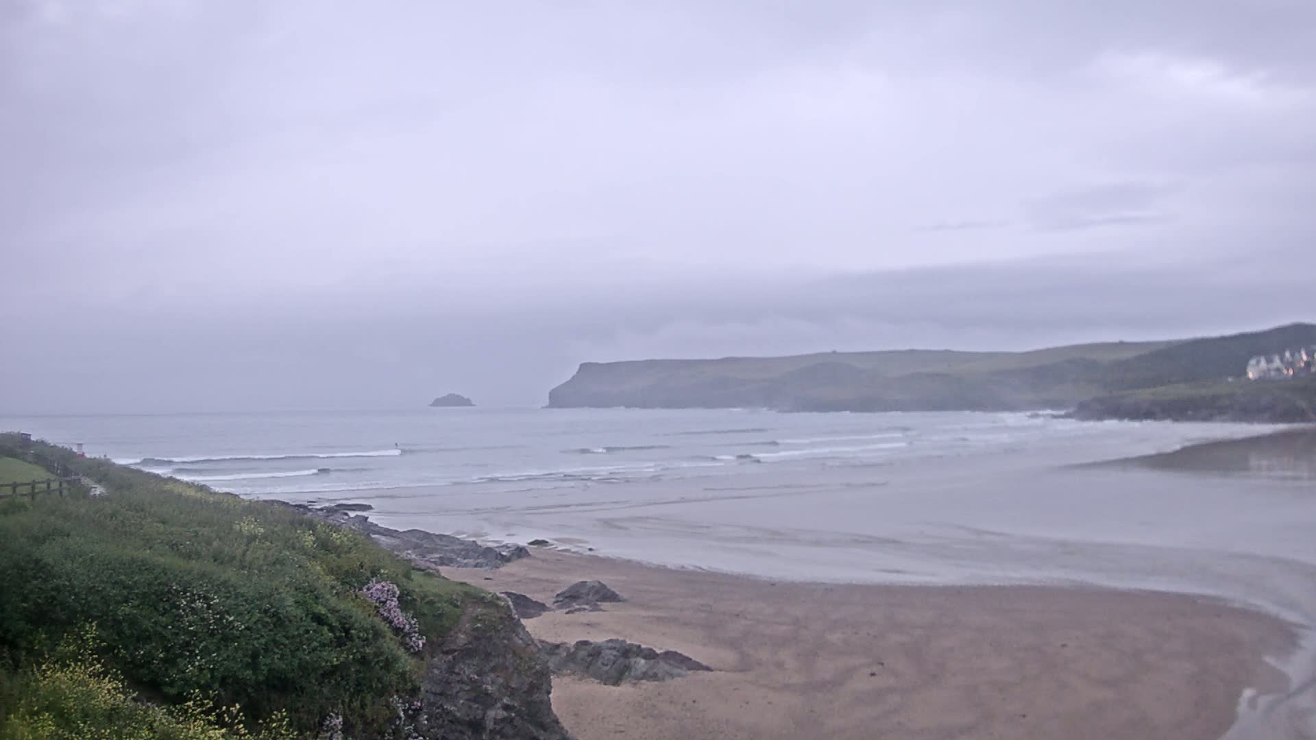 A sandy beach stretches out to a calm sea under an overcast sky, with rolling hills and a small island in the distance.