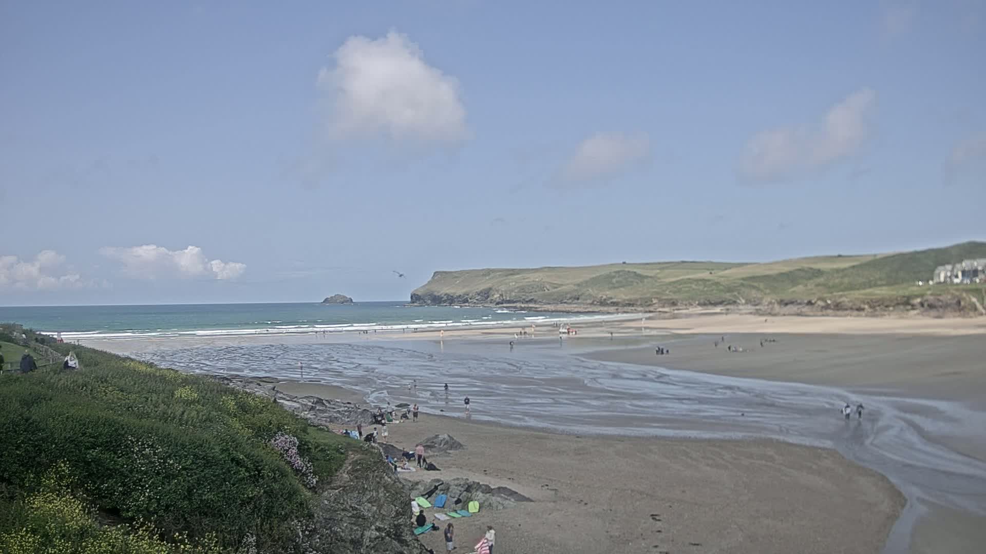 A sandy beach with a few people on it stretches out to a calm ocean under a mostly sunny sky, with a small island visible in the distance.