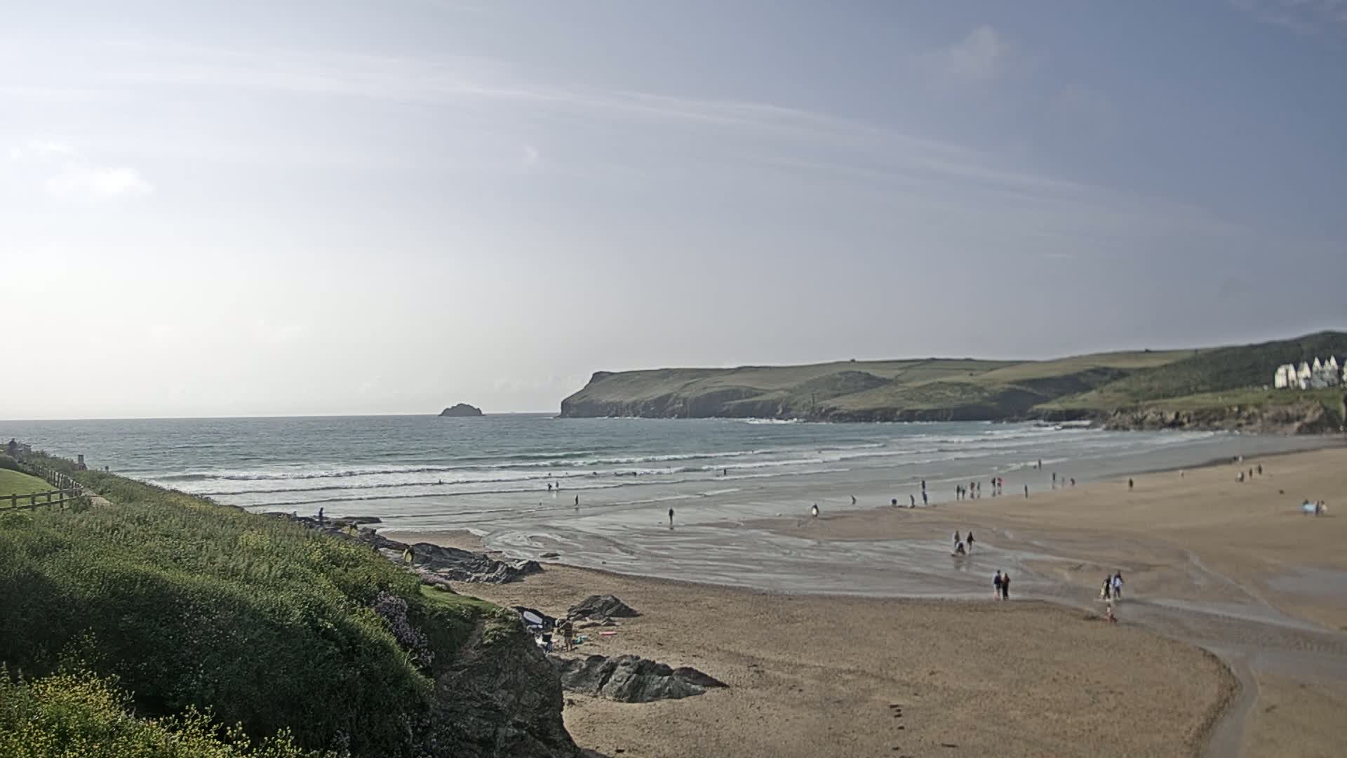 A sandy beach with people on it stretches along a coastline under a mostly clear sky, with gentle waves rolling into the shore.
