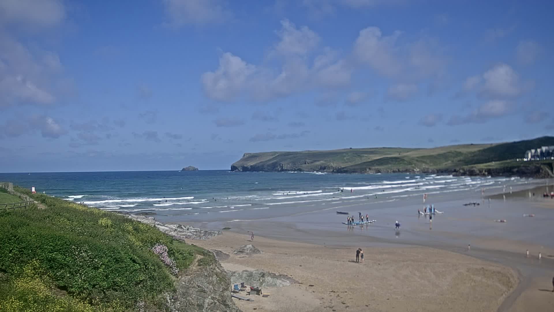 A sandy beach with gentle waves under a partly cloudy blue sky, people enjoying the water and sand.