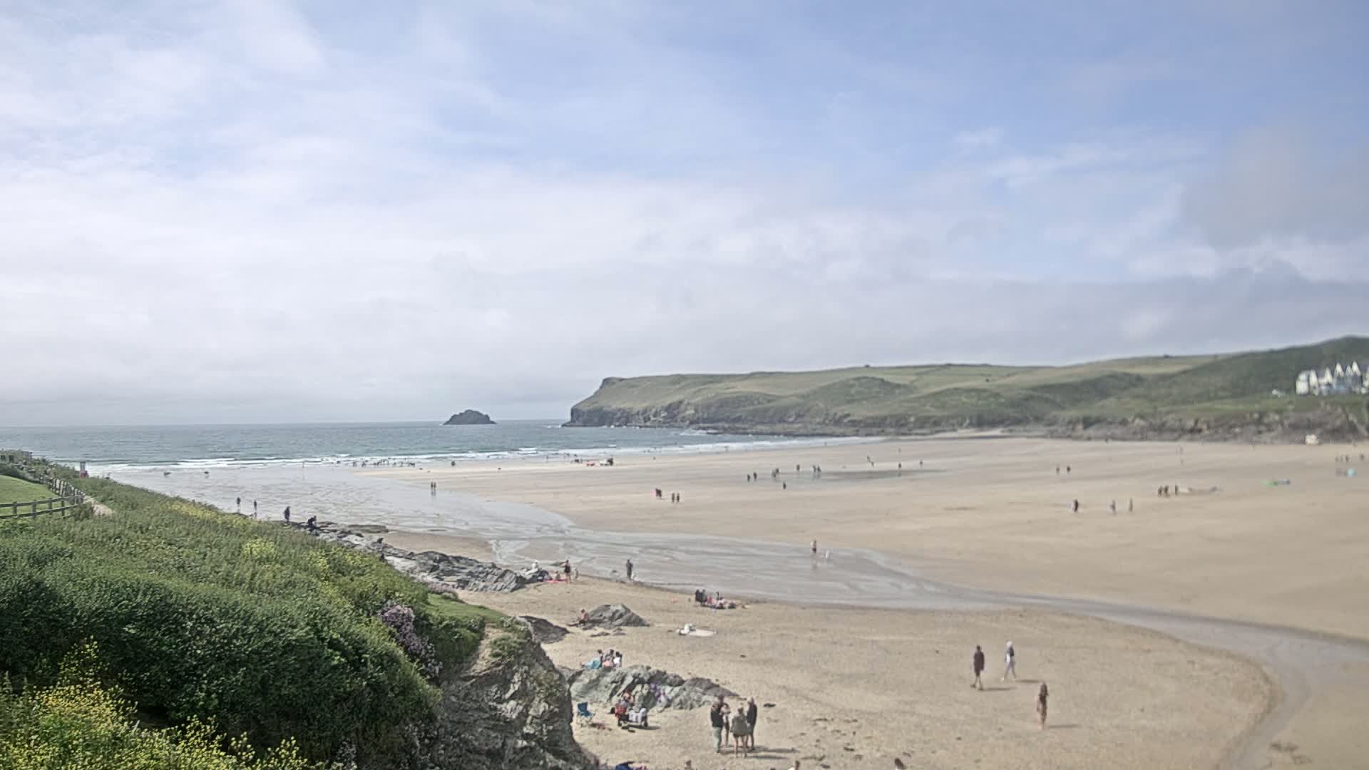 A sandy beach with scattered people stretches out to a calm ocean under a partly cloudy sky, backed by green hills and a small island in the distance.