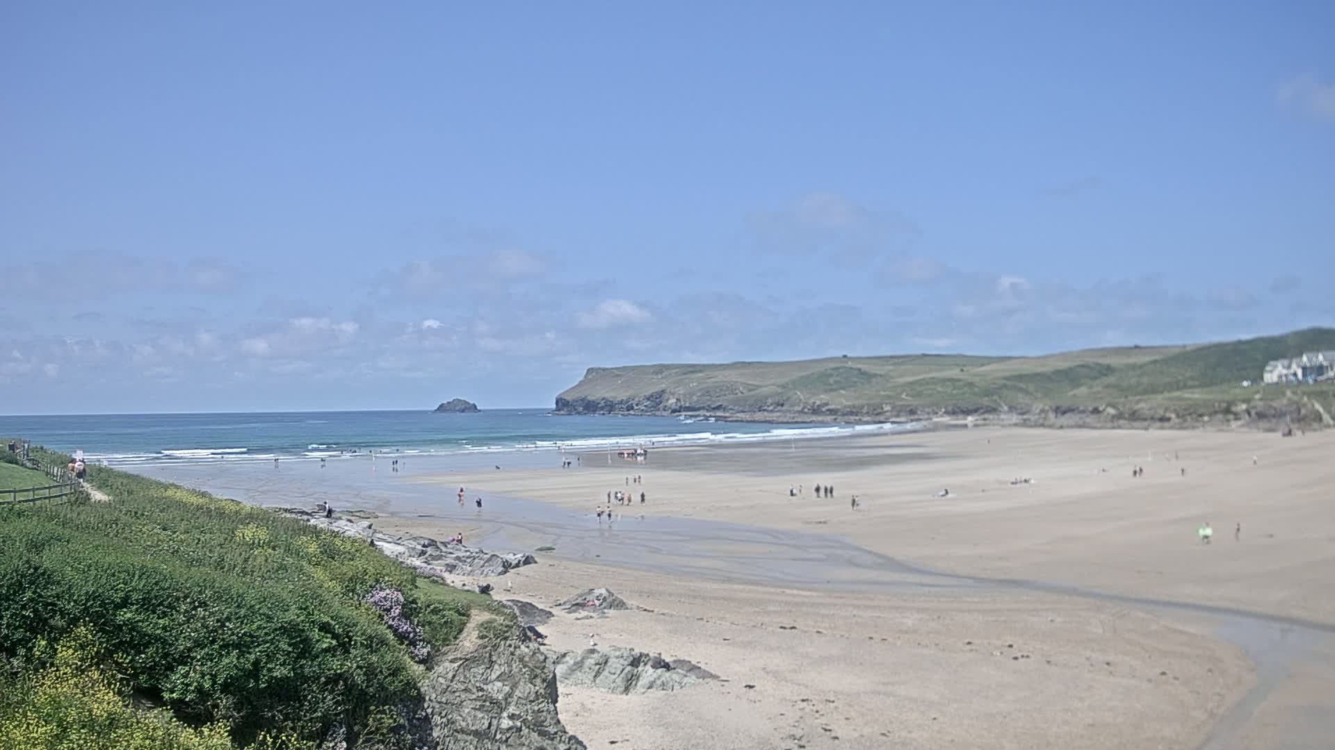 A wide sandy beach with a few people on it, small waves in the ocean, and green hills under a mostly sunny sky.