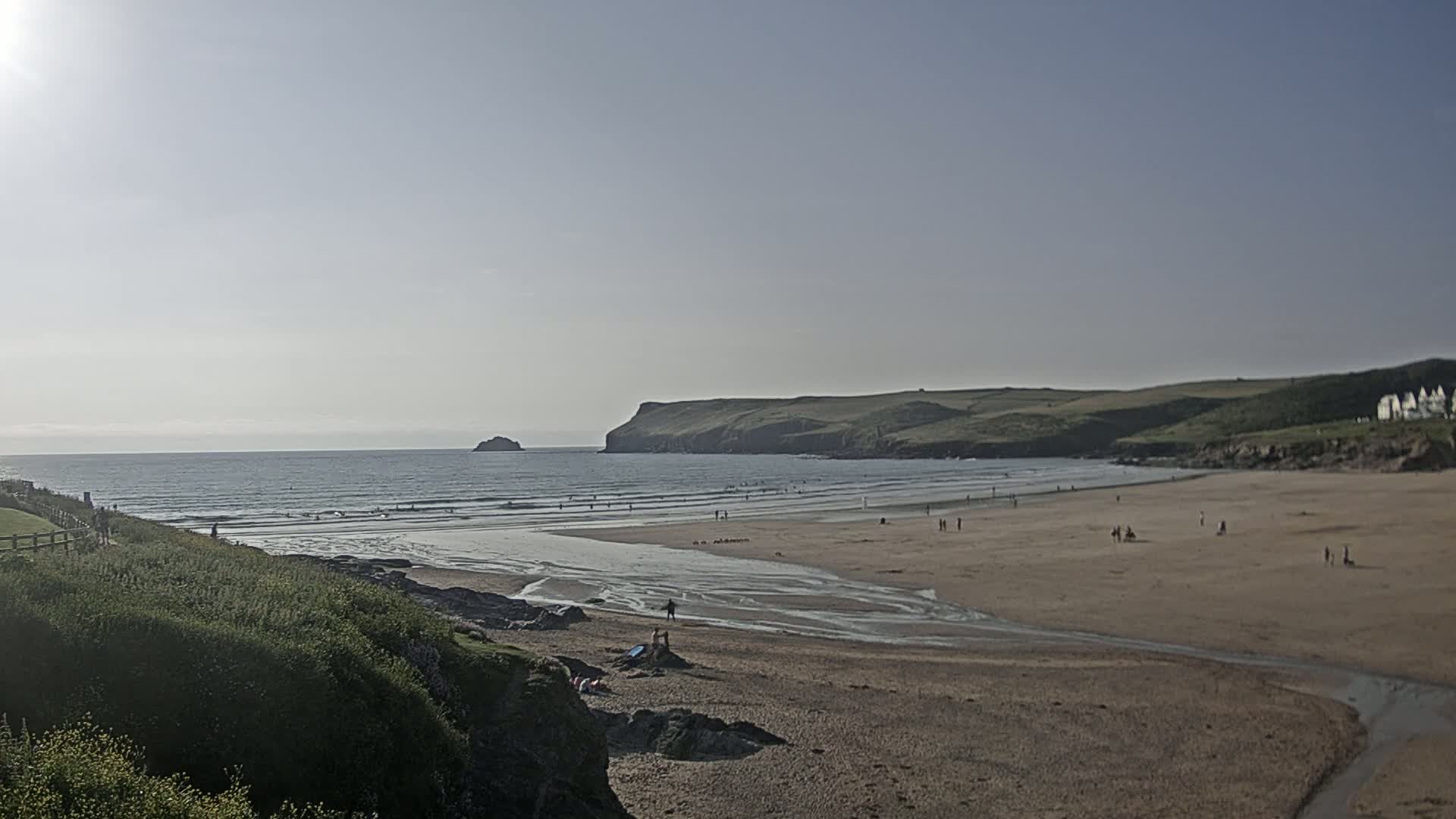 A sandy beach with a calm ocean, a small island in the distance, and a few people scattered across the shore under a bright, sunny sky.