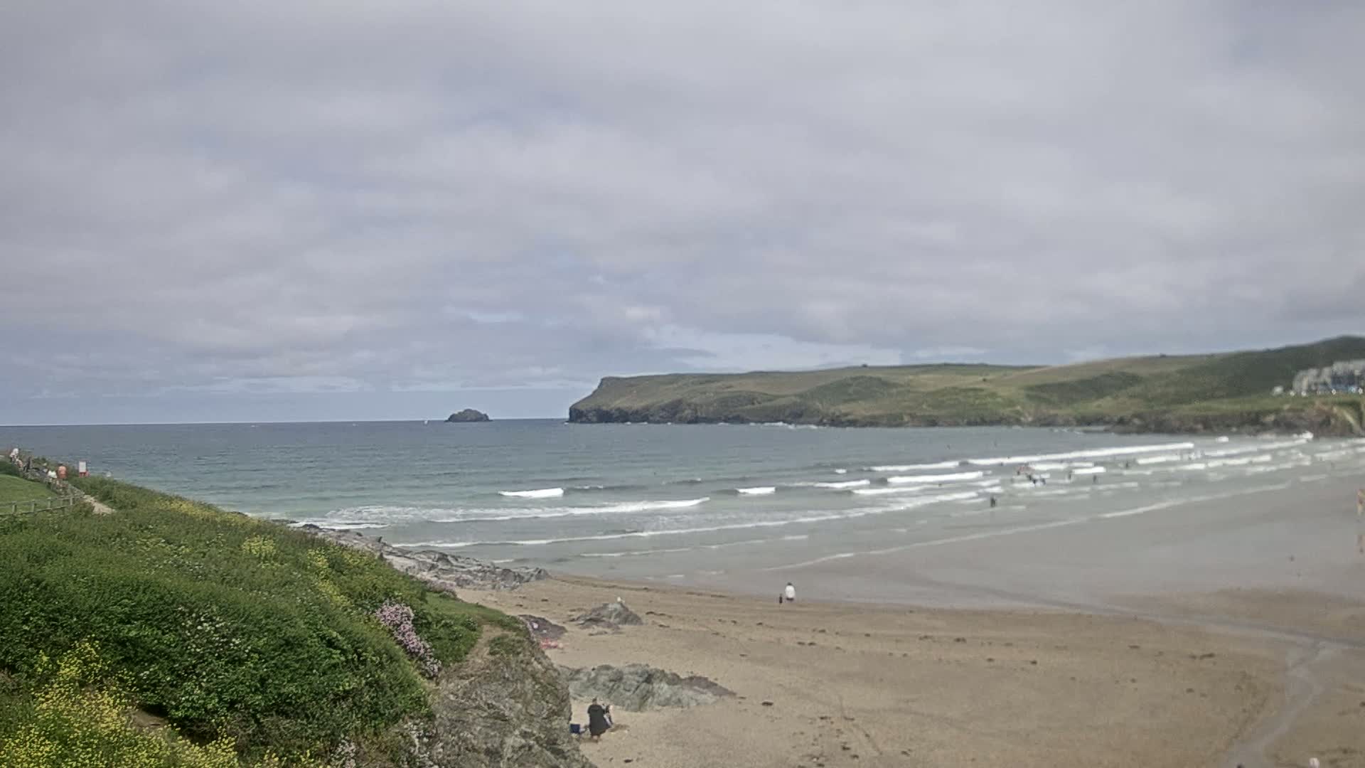 A sandy beach with small waves under a cloudy sky, backed by green hills and a distant rocky island.