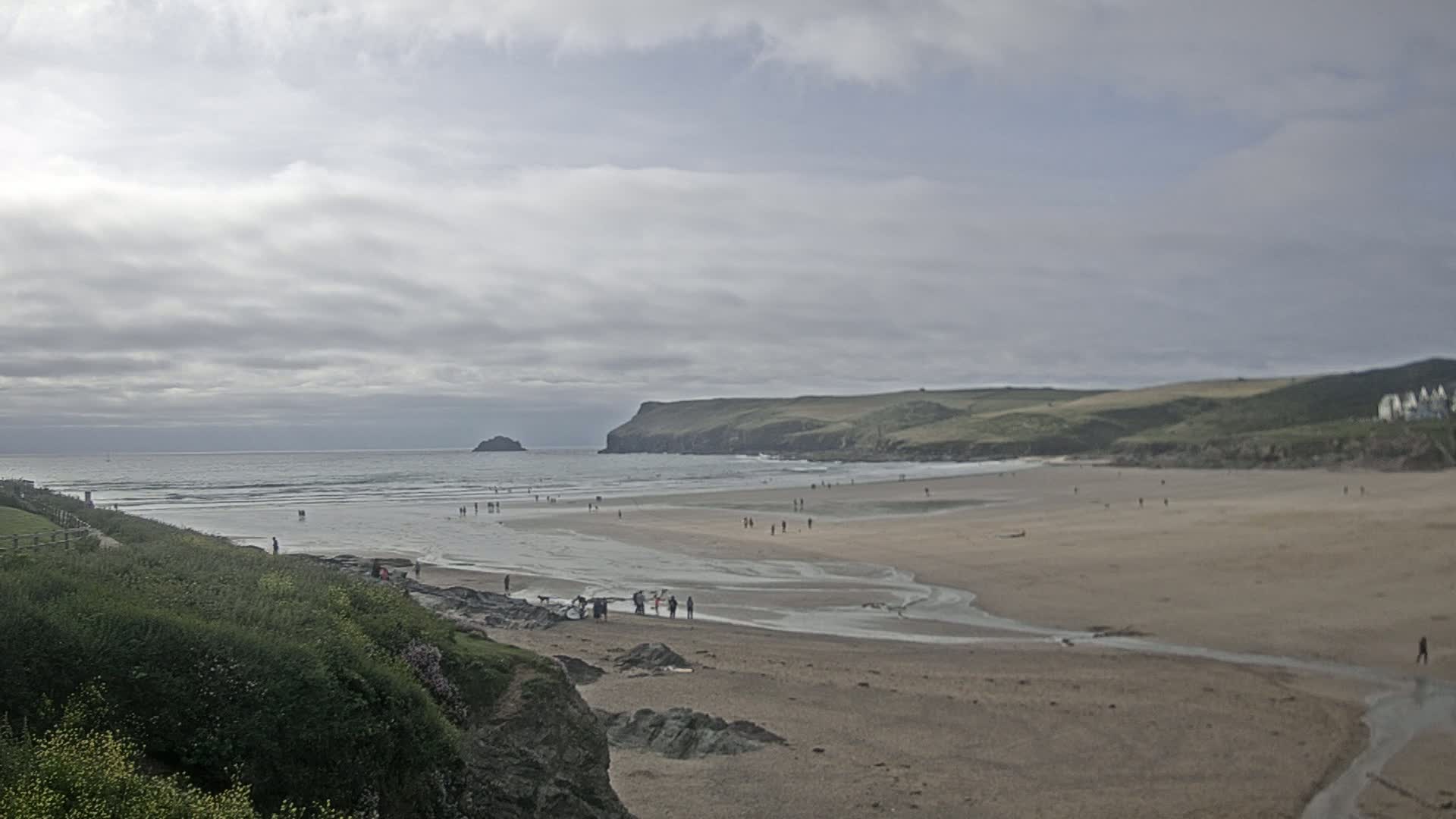 A sandy beach with a few people on it stretches out below a cloudy sky, with a small island and a green hill visible in the distance.