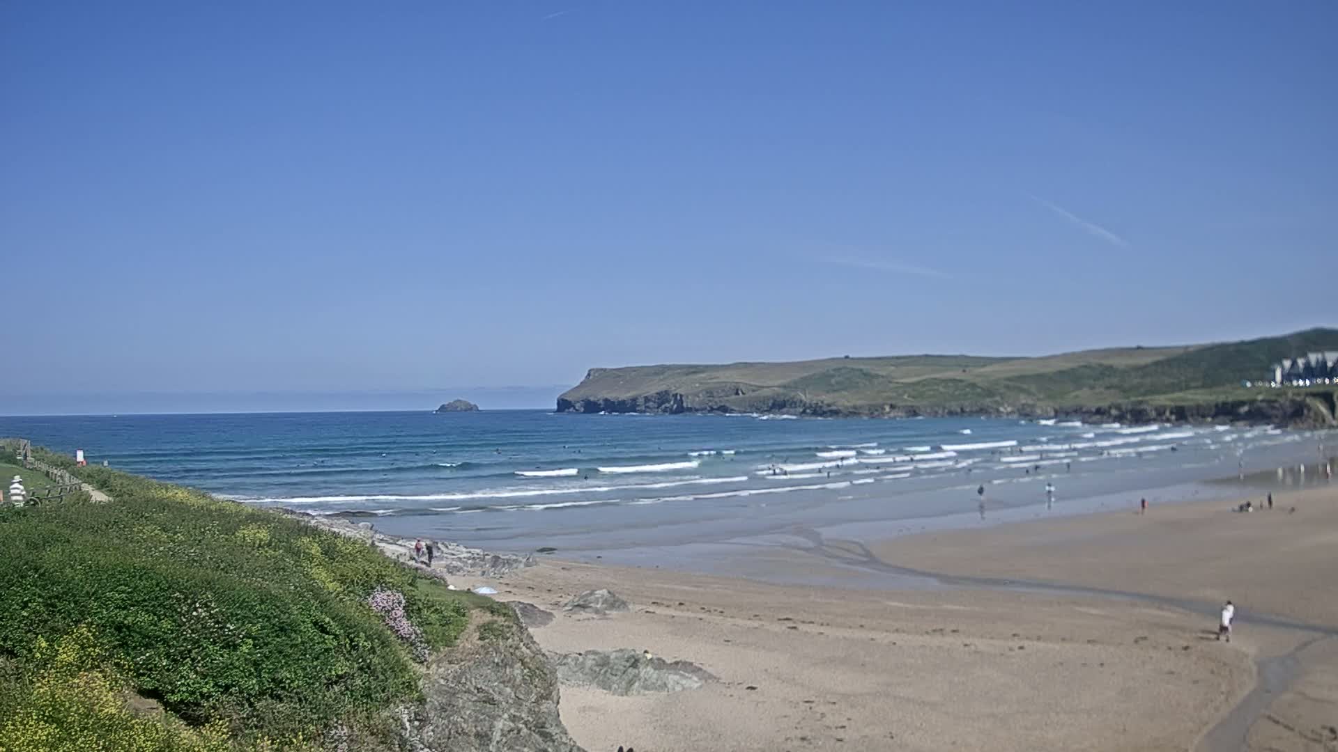 A sandy beach with gentle waves under a clear blue sky, with people scattered along the shore and in the water.