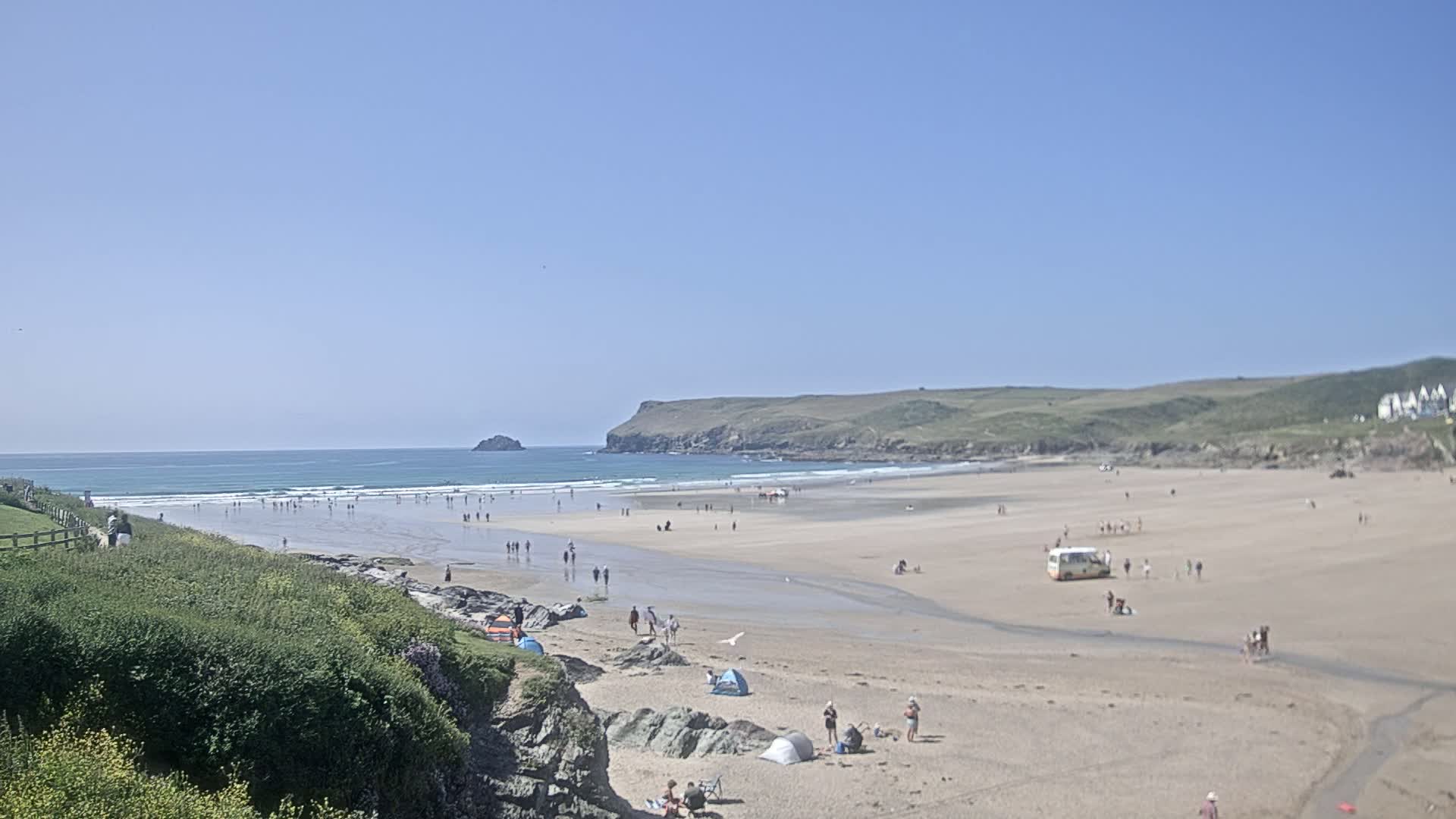 A sandy beach with many people on it and a few small tents, under a clear blue sky, stretches to a calm ocean with small waves.