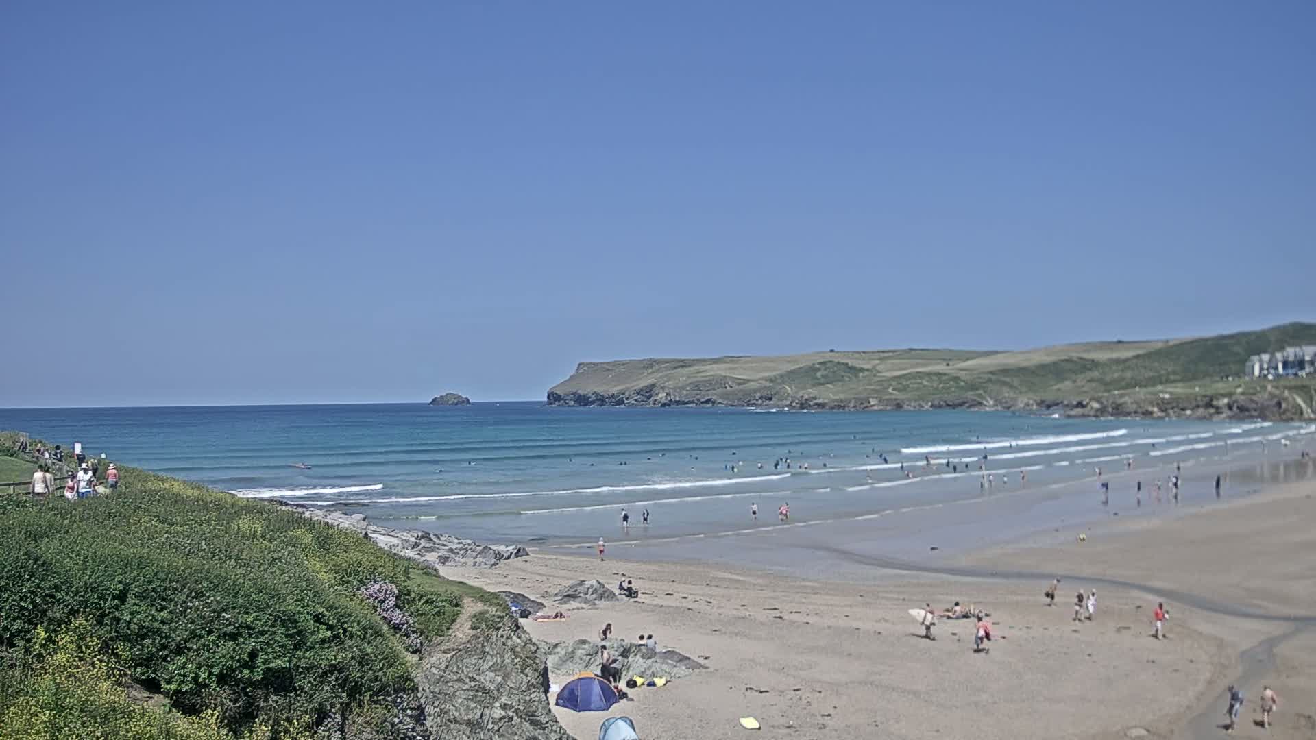 A sandy beach with many people swimming and sunbathing stretches out under a clear blue sky, small waves lapping the shore.