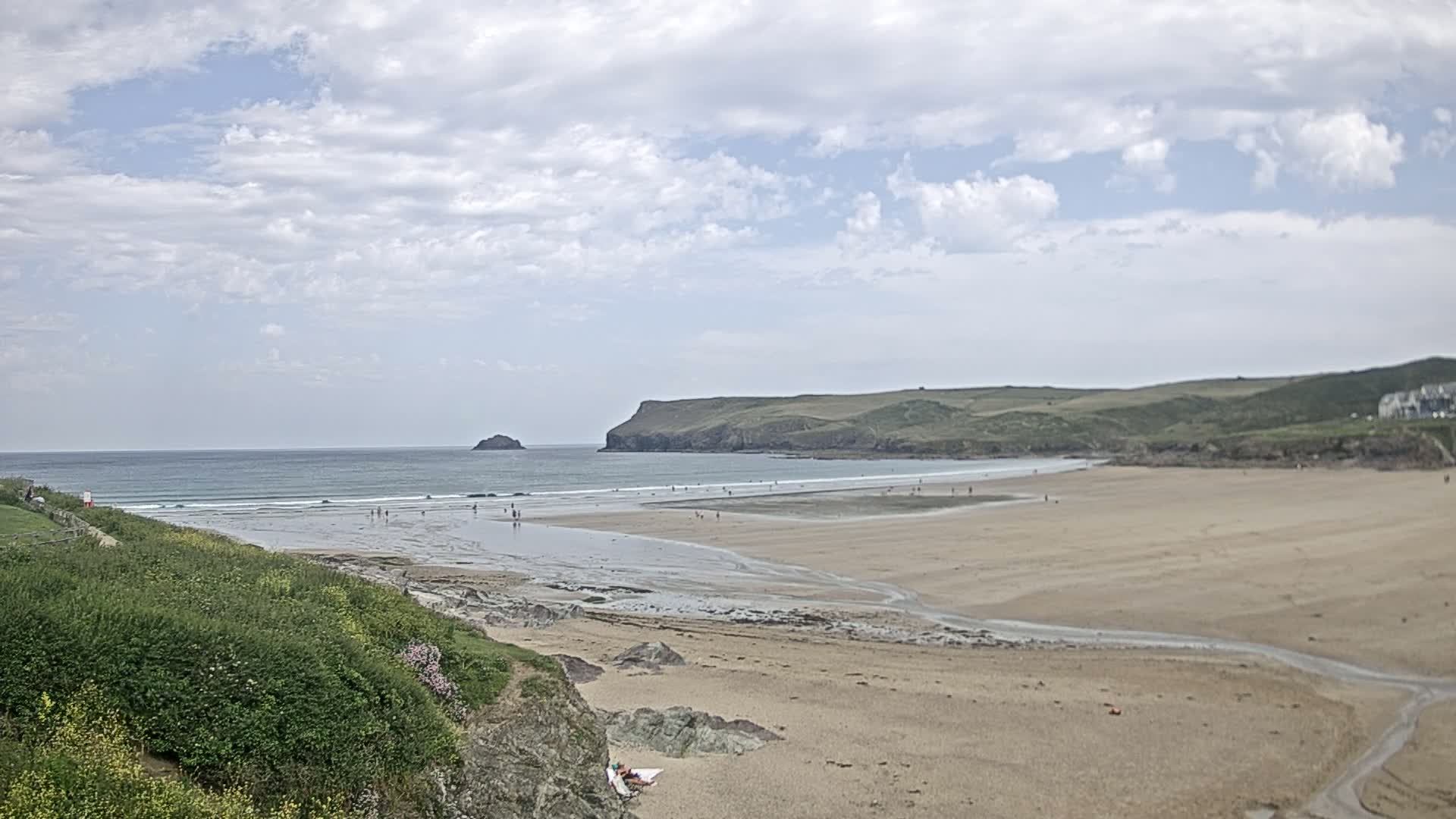 A sandy beach stretches out to a calm ocean under a partly cloudy sky, with people scattered along the shoreline and a small island visible in the distance.