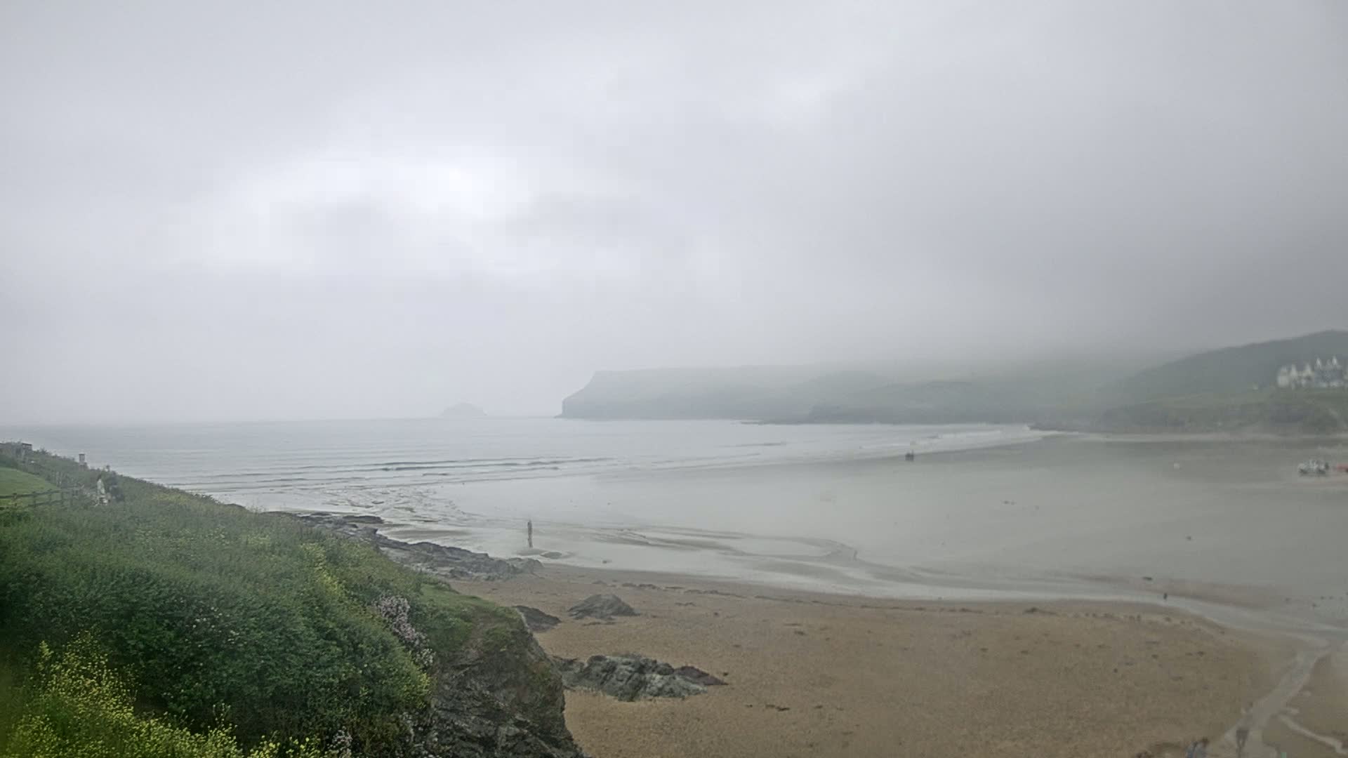 A foggy coastal scene shows a sandy beach meeting a calm, grey ocean, backed by low, hazy hills.