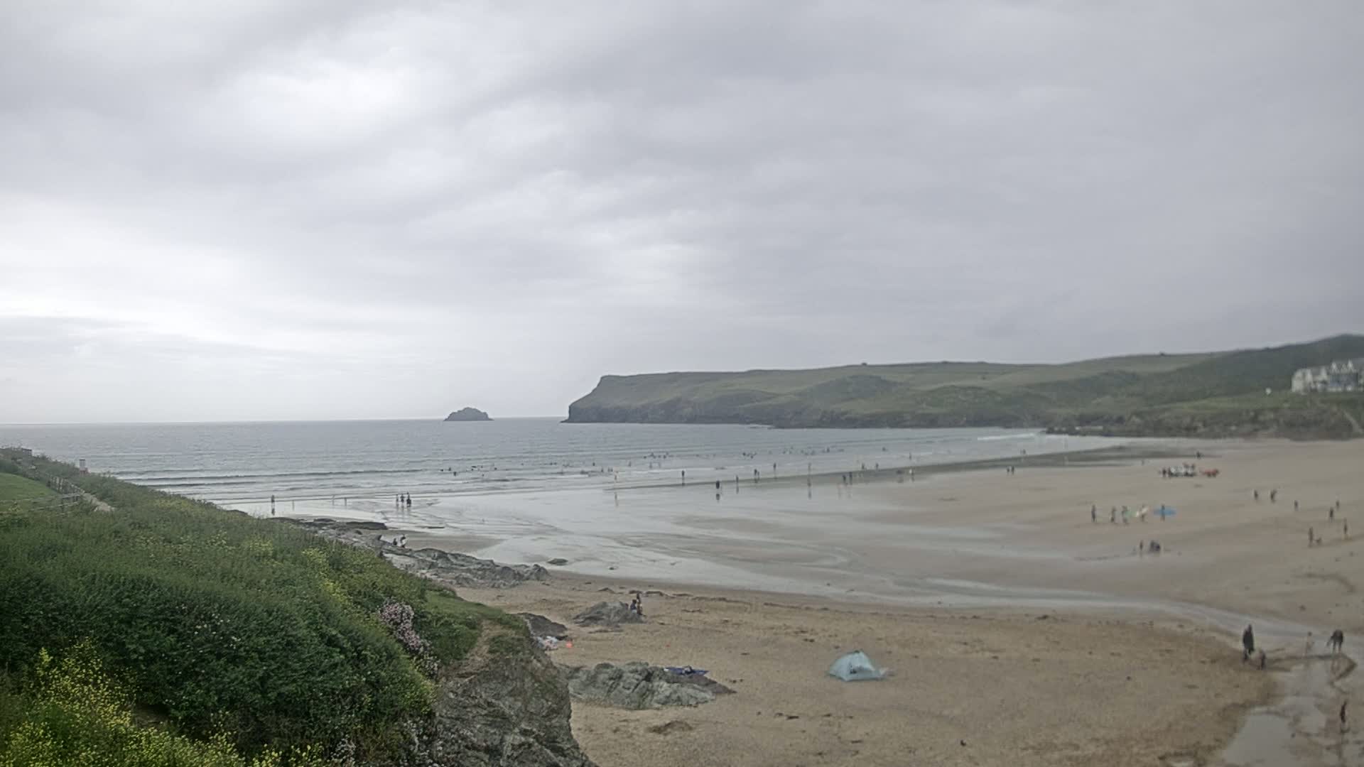 A sandy beach with a few people on it stretches between a grassy cliff and the ocean under an overcast sky.