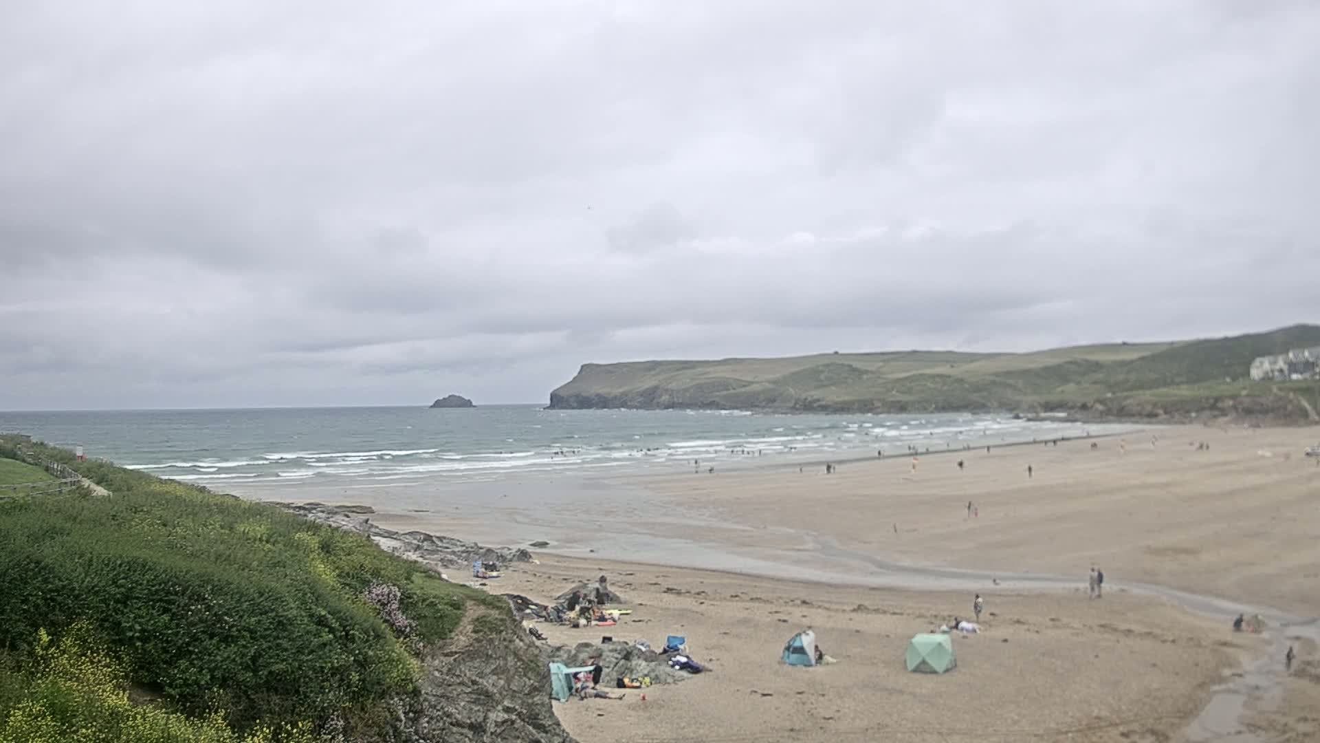 A sandy beach with people scattered across it, under a cloudy sky, features gentle waves rolling into shore with a small island visible in the distance.