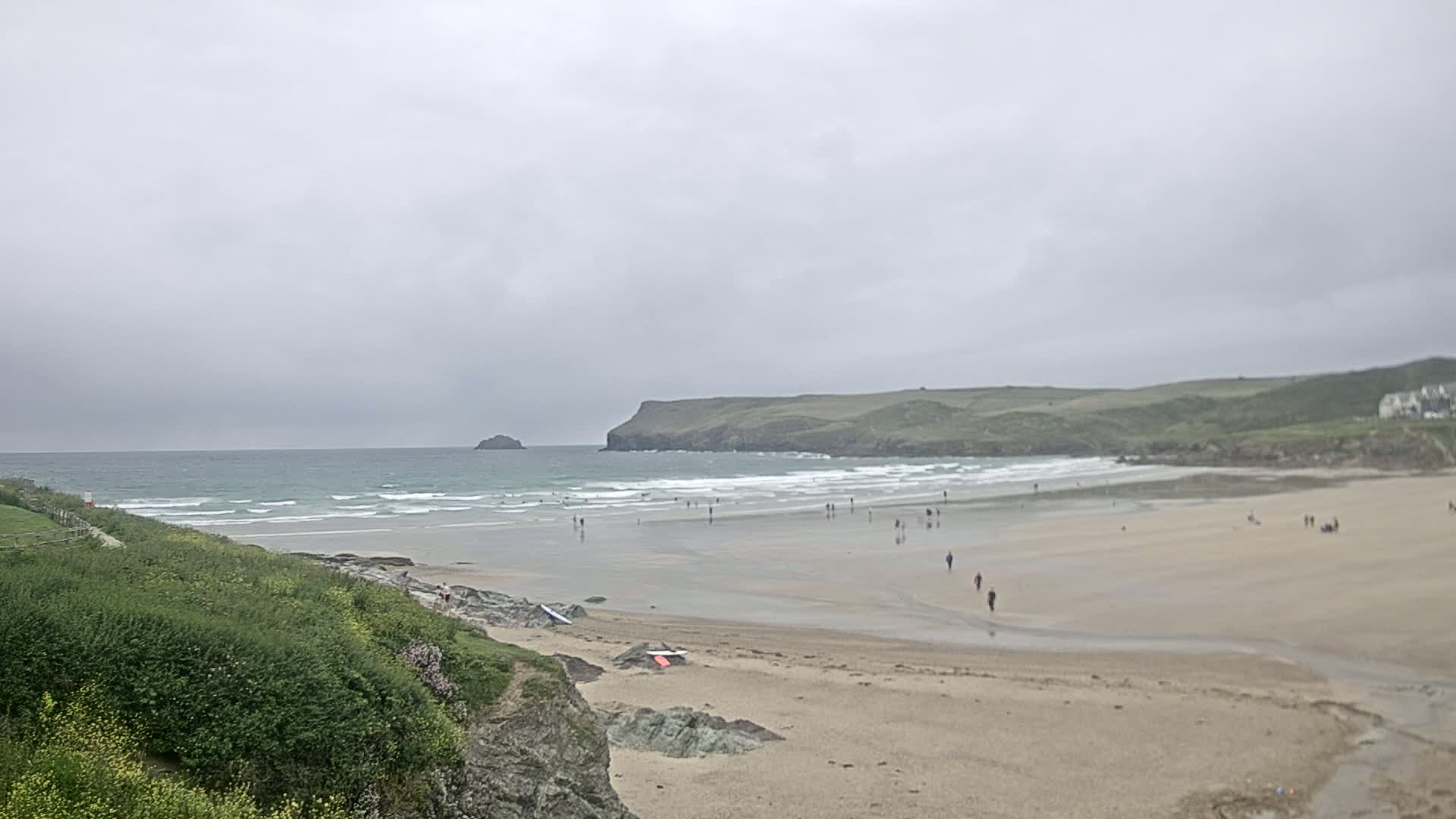 A wide sandy beach with small waves under an overcast sky, featuring a few people walking and standing on the sand and in the water.