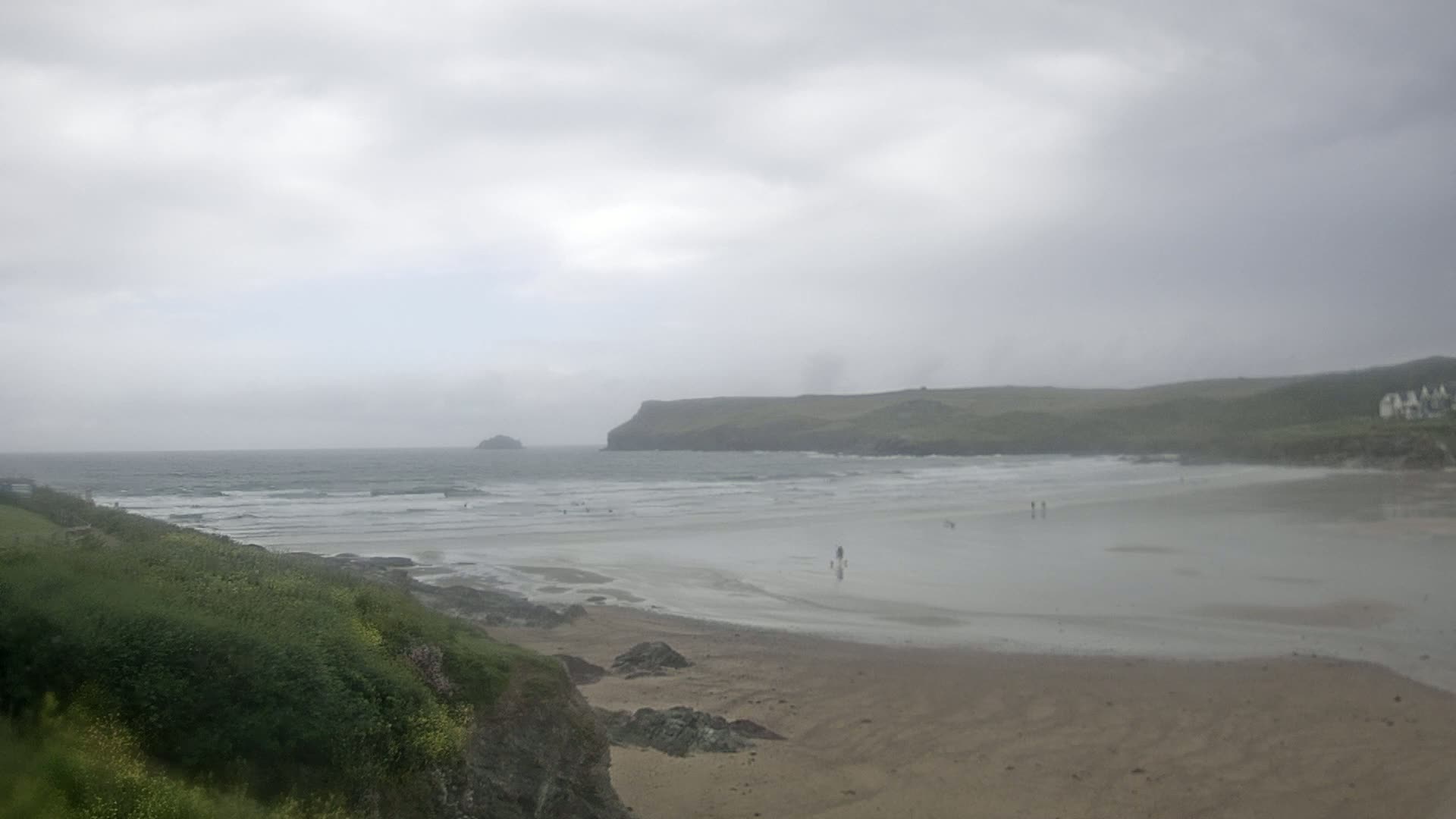 A sandy beach with small waves and a few people stretches out under an overcast sky, backed by green hills and a distant island.