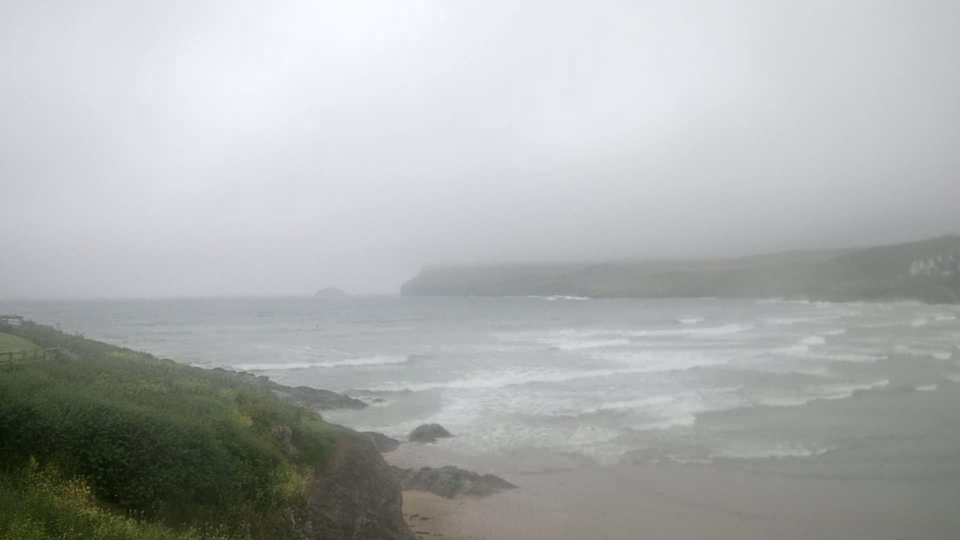 A foggy, overcast day reveals a choppy sea meeting a sandy beach bordered by green vegetation and distant, shrouded headlands.
