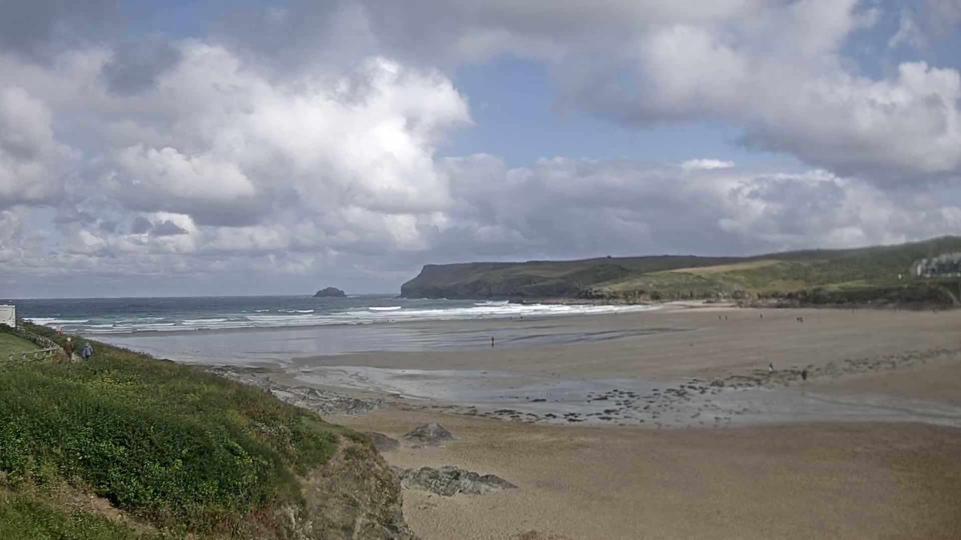 A sandy beach stretches out to a wavy ocean under a partly cloudy sky, with green hills and a distant island in the background.