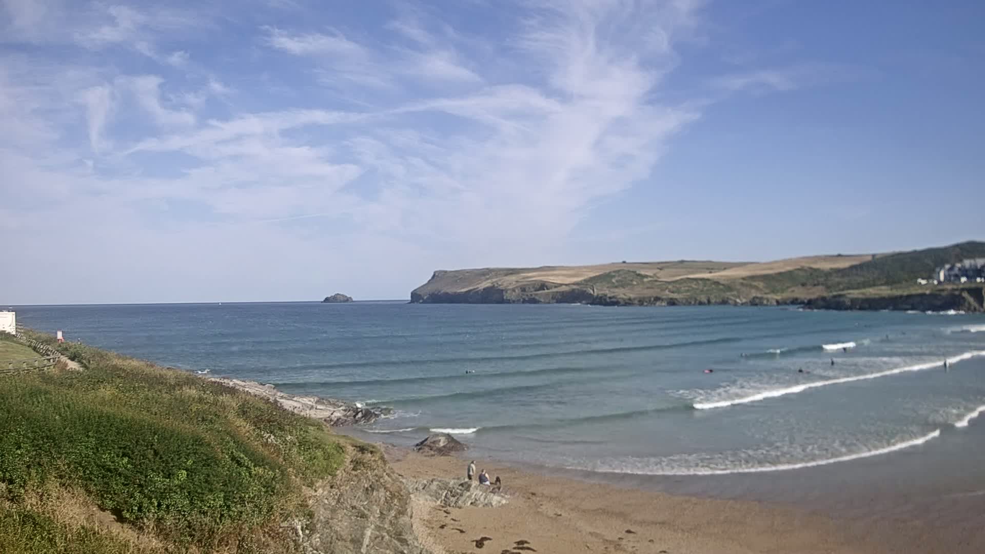 A sandy beach with gentle waves and a few surfers under a mostly sunny sky.