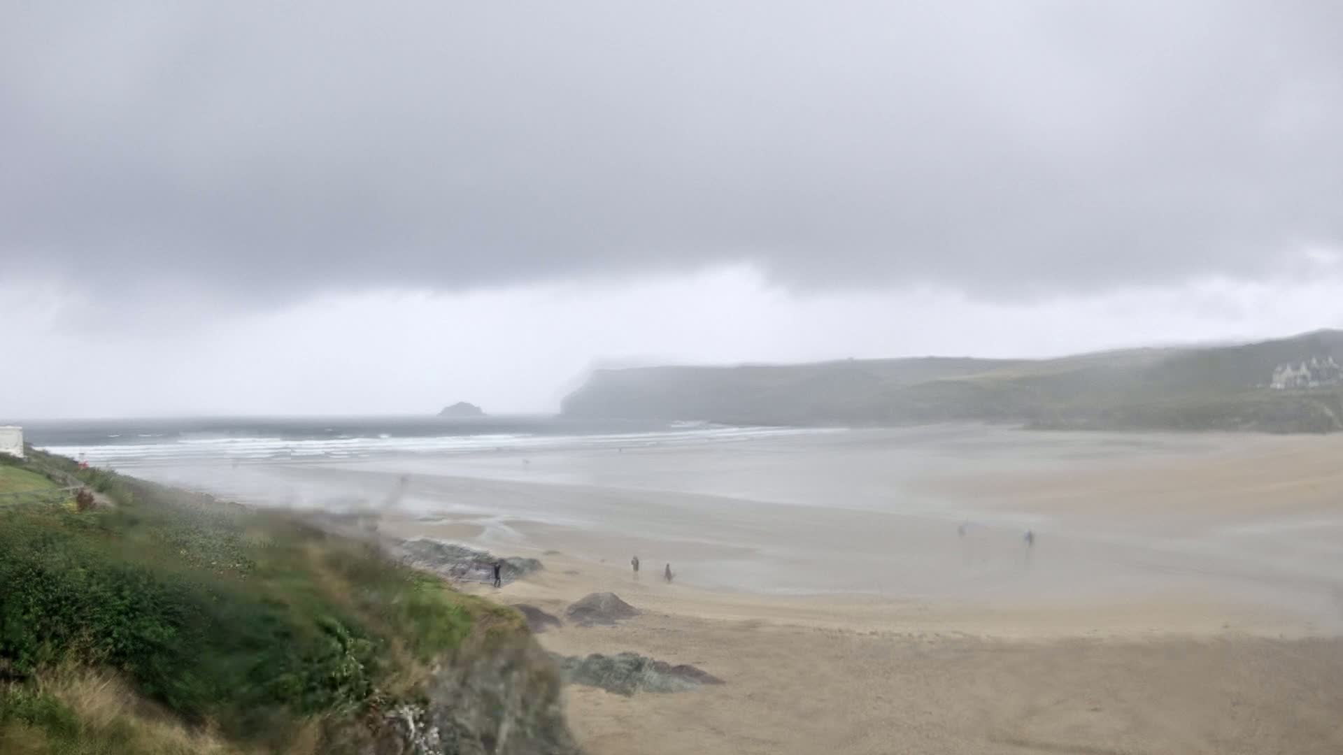 A wide, sandy beach stretches under a cloudy, overcast sky, with a few people visible in the distance and waves breaking in the ocean beyond.
