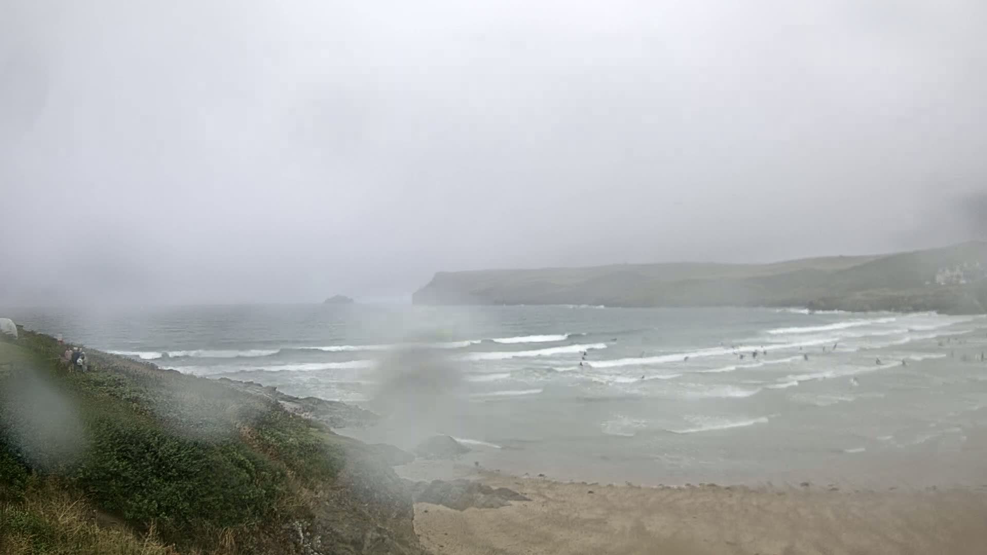 A rain-blurred view of a beach with small waves and several surfers, seen from a grassy clifftop on a hazy, overcast day.