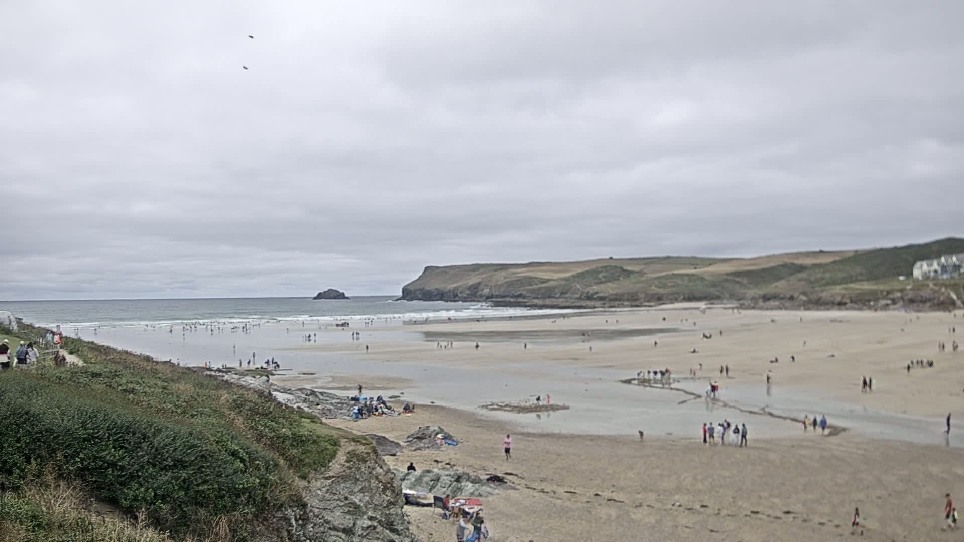 A wide sandy beach with many people on it, under an overcast sky, is bordered by green hills and a calm ocean with small waves.