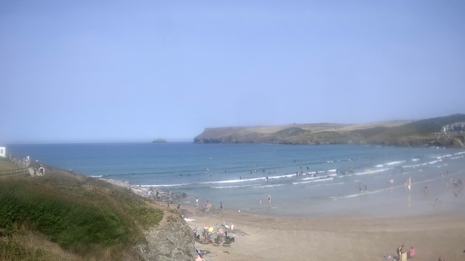 A wide sandy beach with many people on it, under an overcast sky, is bordered by green hills and a calm ocean with small waves.