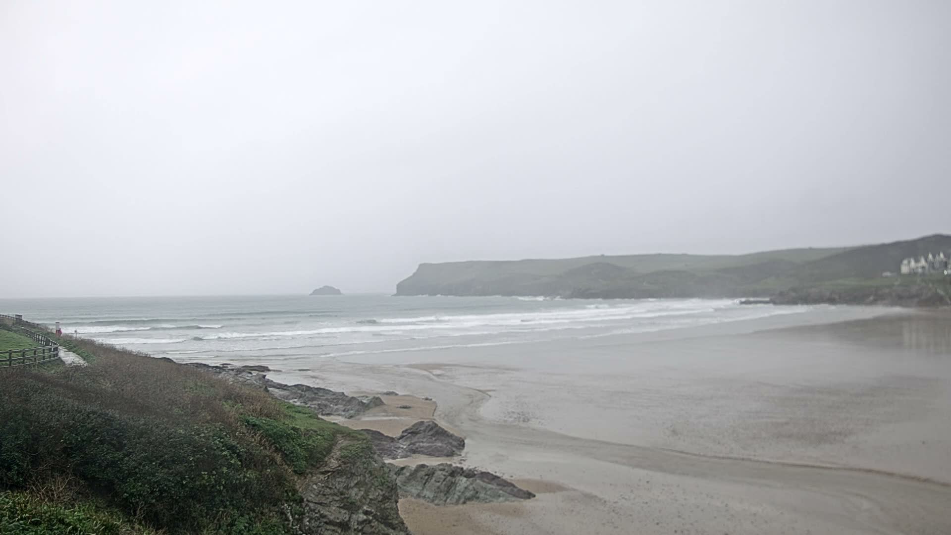 A vast, grey sandy beach with gentle waves crashing onto the shore is bordered by a green, rocky cliff on the left and a distant headland with houses on the right, all under a heavily overcast and muted grey sky.