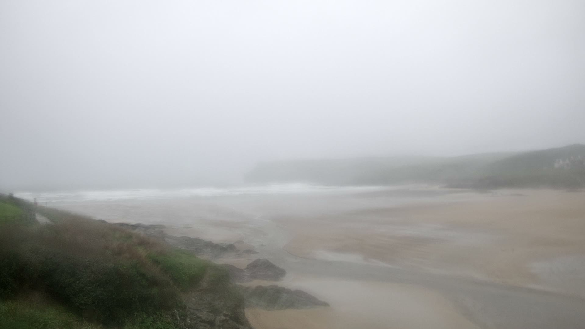 A desolate coastal landscape is enveloped in dense fog, revealing waves breaking on a sandy beach bordered by a rocky, grassy shoreline, with faint hills and distant structures barely discernible through the gloom.
