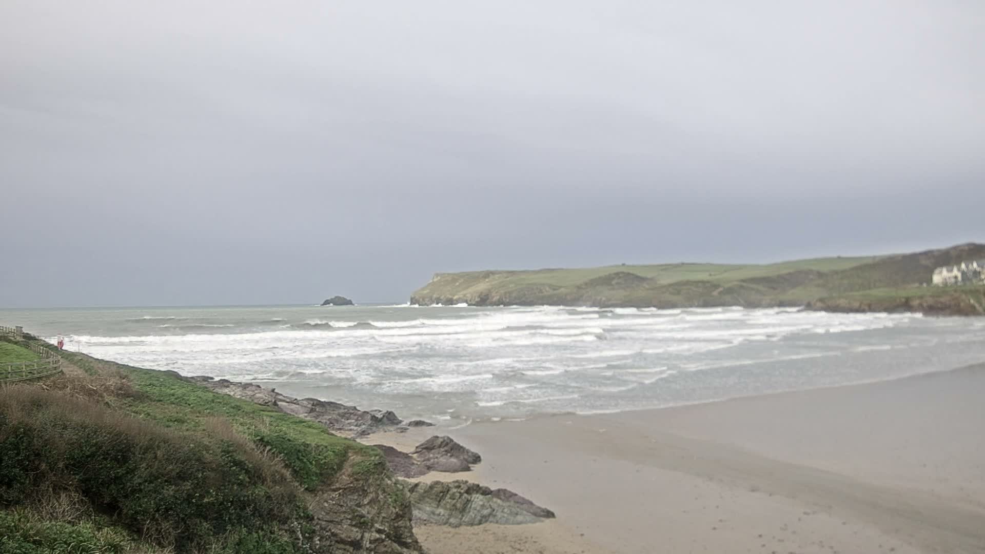 Under a uniformly overcast sky, an expansive, grey sandy beach with gentle waves stretches towards a distant rocky islet and green headlands, with a vegetated embankment and path visible on the left.