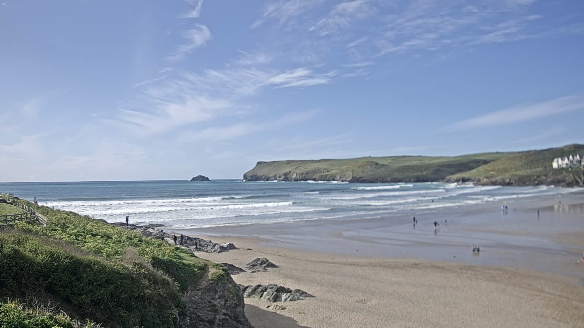 Under a uniformly overcast sky, an expansive, grey sandy beach with gentle waves stretches towards a distant rocky islet and green headlands, with a vegetated embankment and path visible on the left.