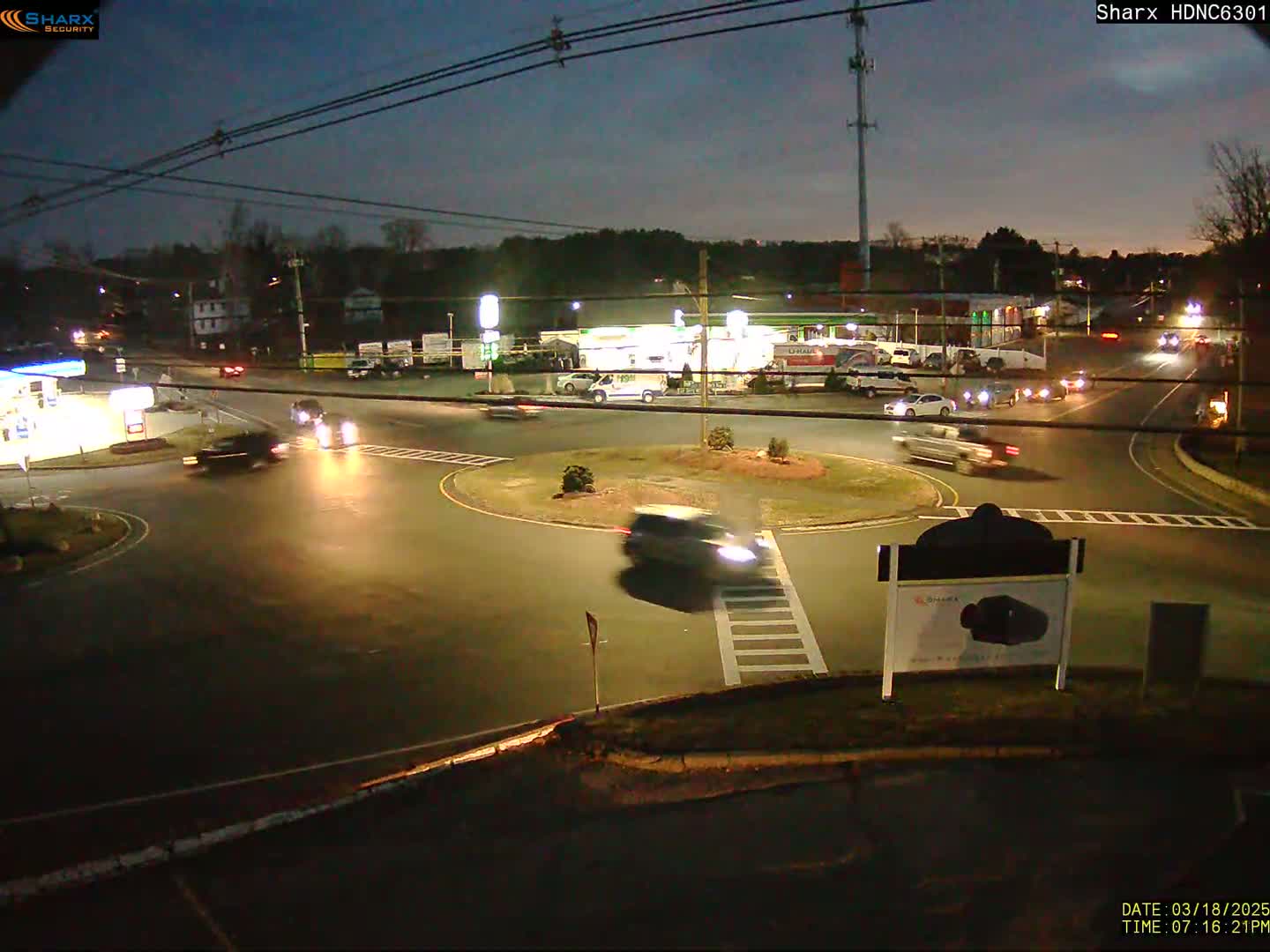 A nighttime view of a roundabout intersection with several cars and businesses visible under a dark sky.