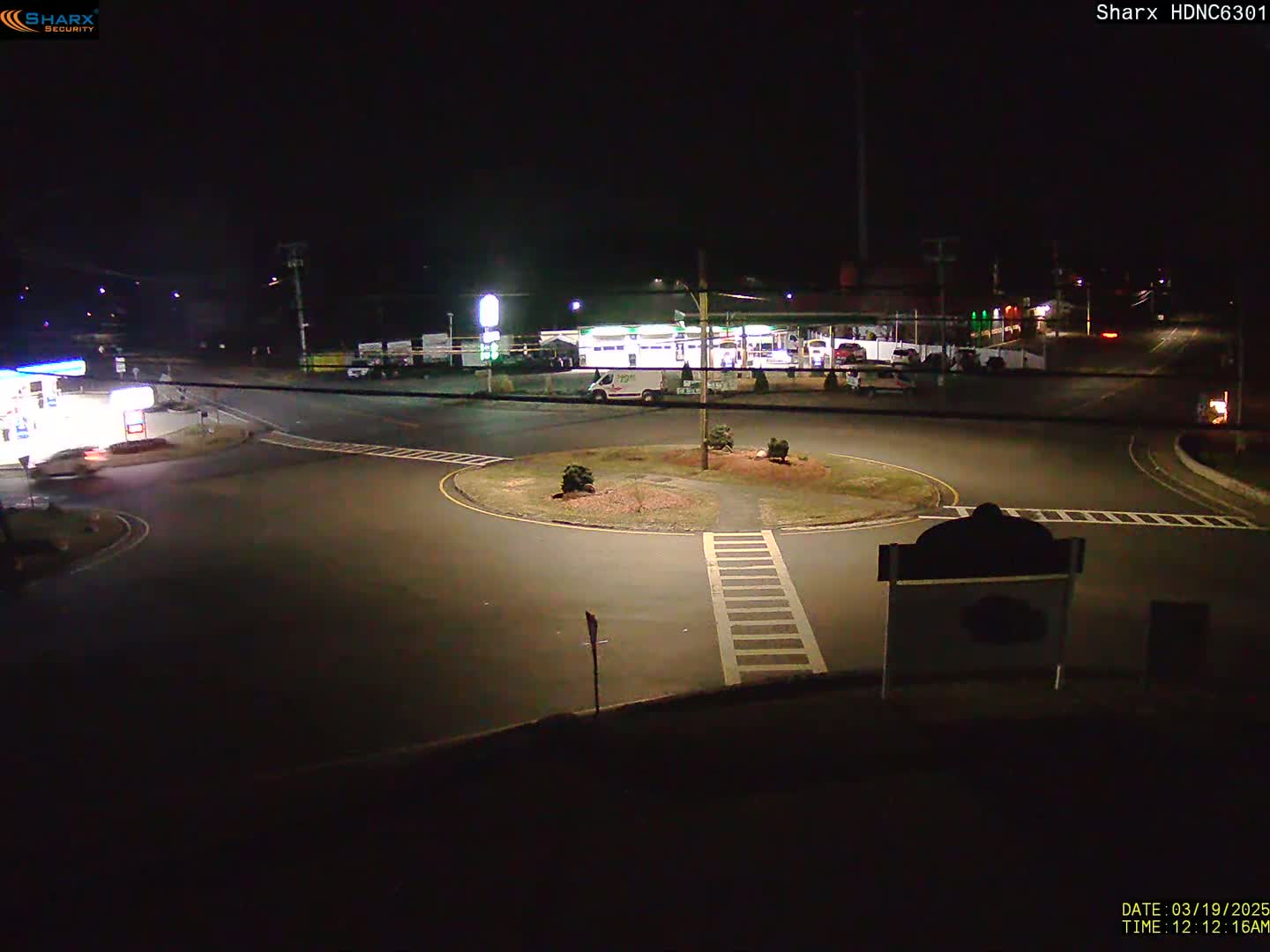 A nighttime view of a roundabout intersection with a gas station and other businesses visible in the background under dark, clear skies.