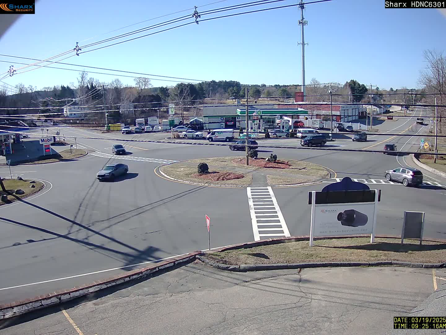 A sunny day shows a traffic circle with several cars, a gas station, and other buildings in the background.