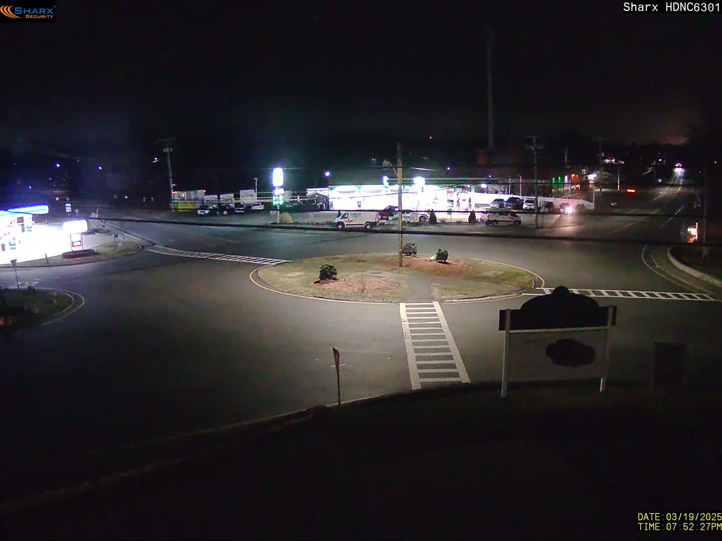 A nighttime view of a roundabout intersection with a gas station and several vehicles visible under dark, clear skies.