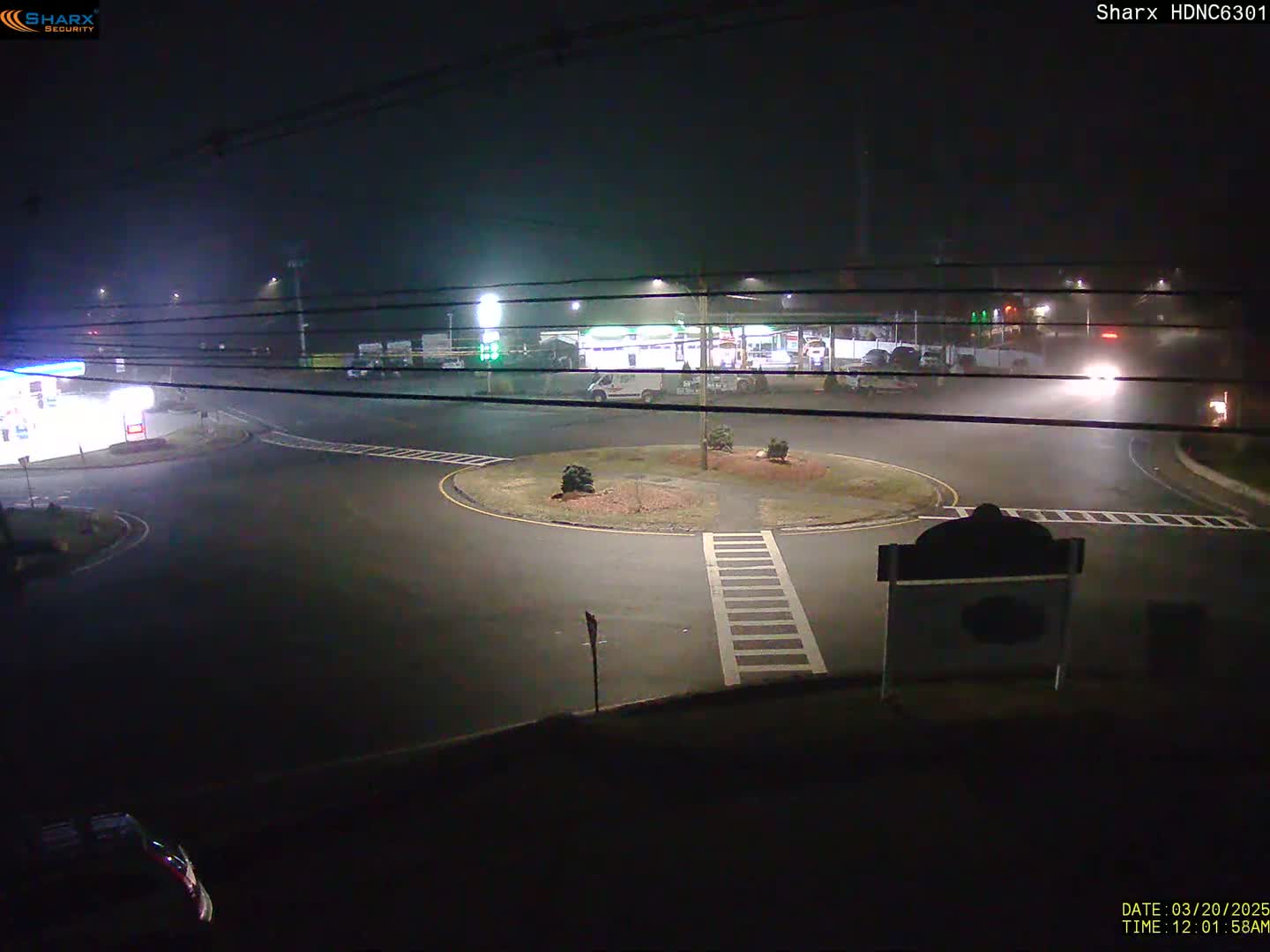 A foggy night scene shows a circular intersection with a gas station and other buildings visible in the background.