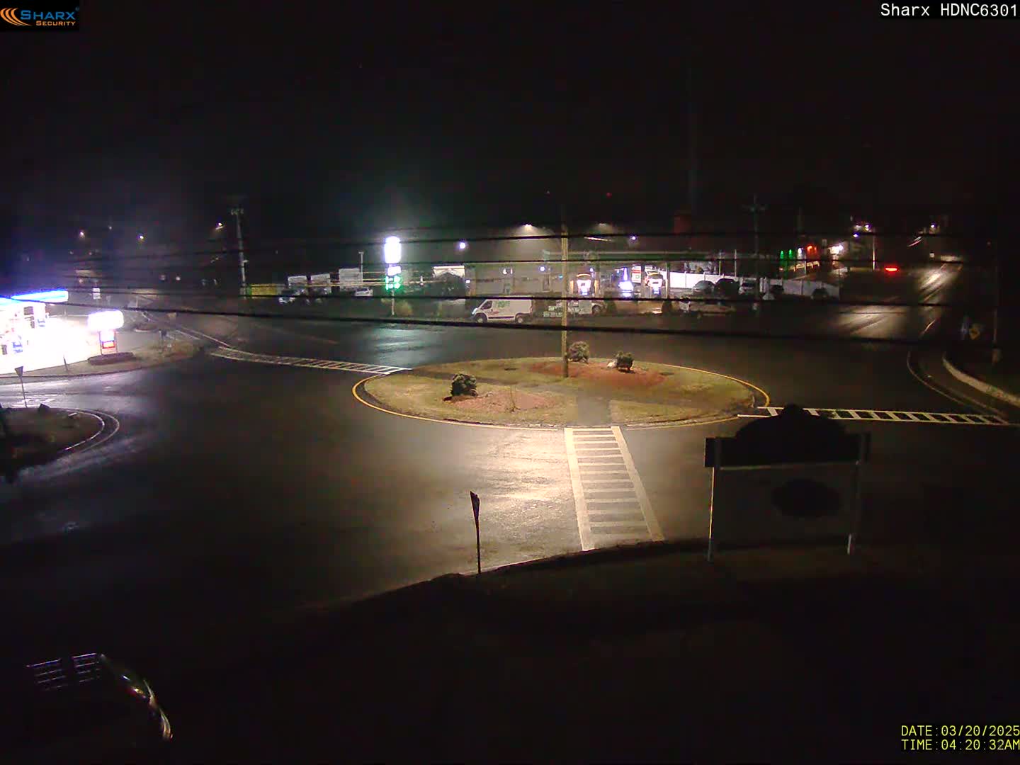 A wet, nighttime view of a roundabout with a gas station and other buildings in the background.