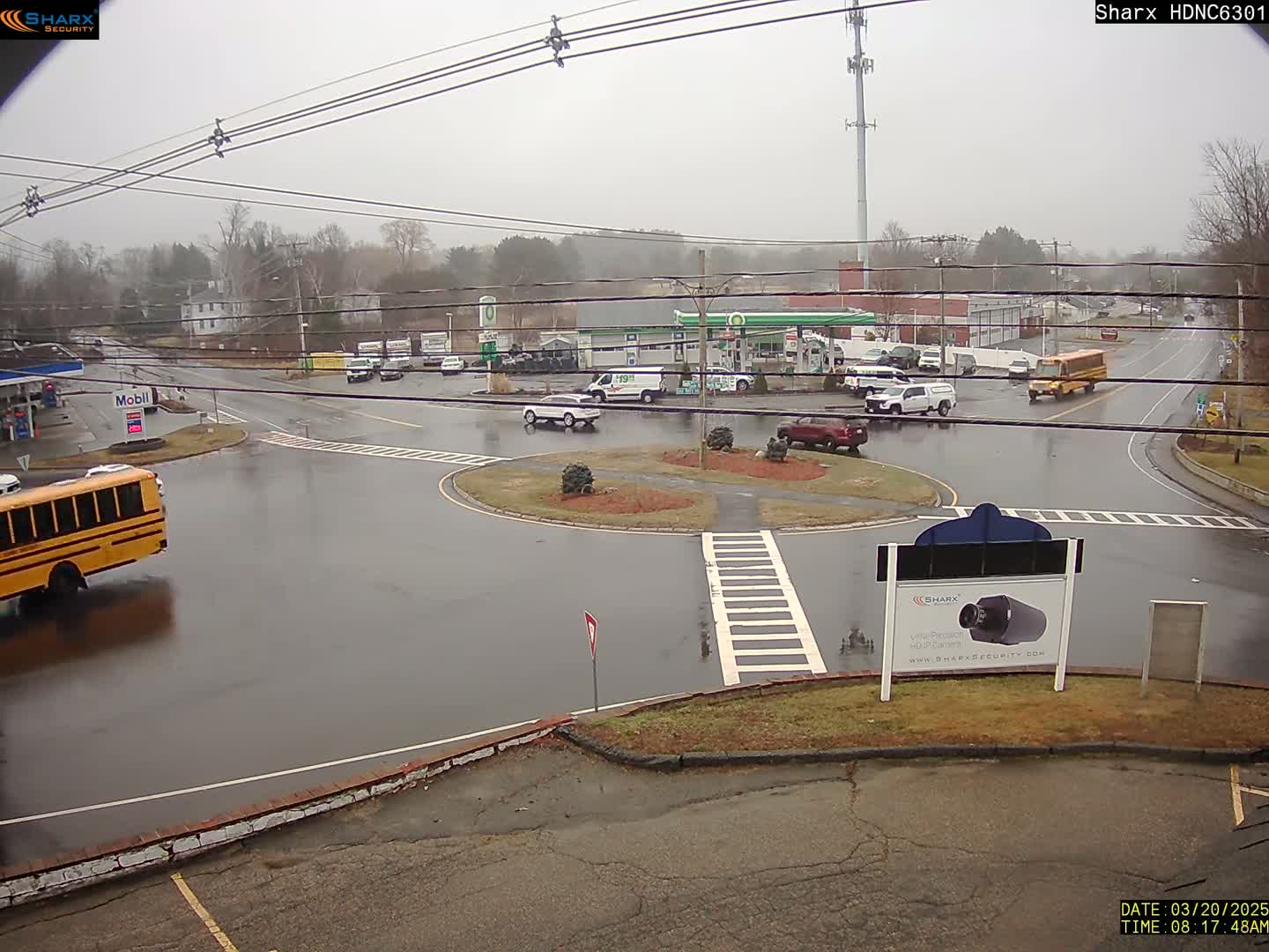 A wet, overcast day reveals a traffic circle with several vehicles, including a school bus, near gas stations and other buildings.