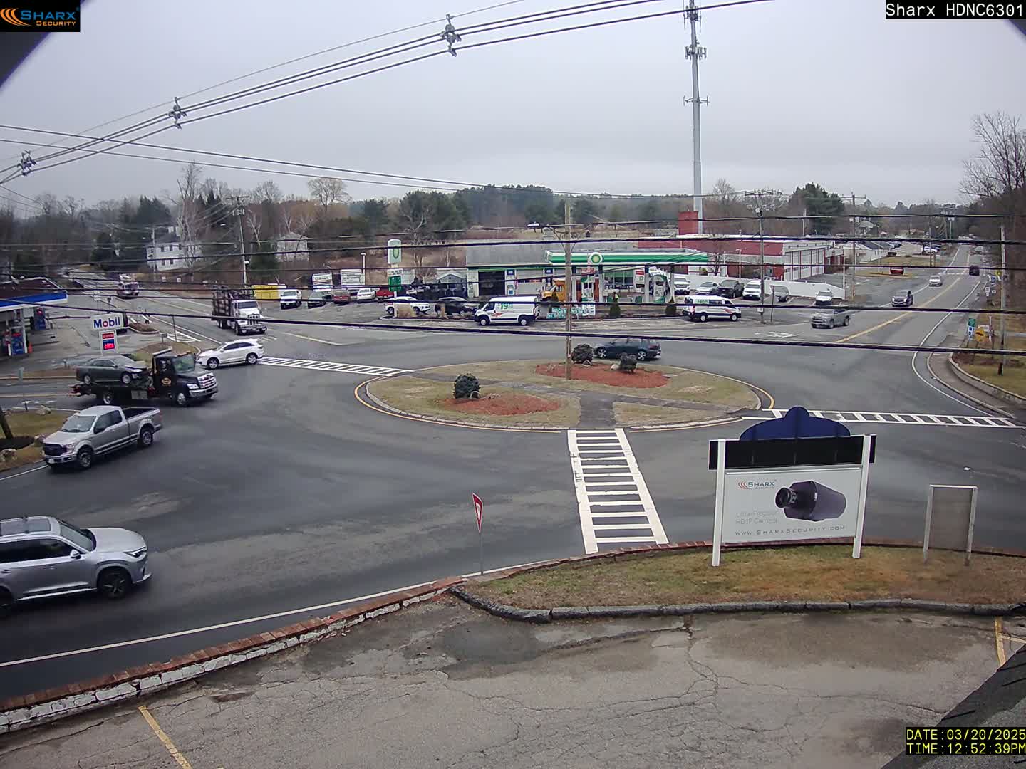 A mostly overcast day shows a view of a highway intersection with a rotary, several cars, and trucks, including a tow truck,  and gas stations nearby.