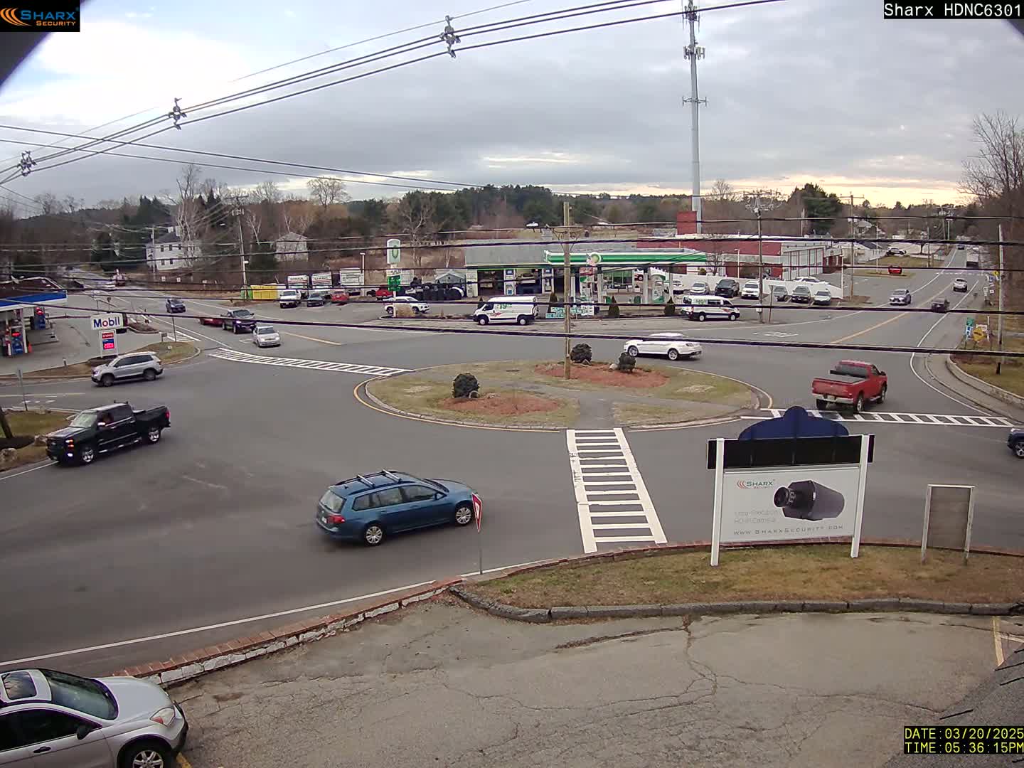 A mostly cloudy day reveals a roundabout intersection with several cars and trucks, including a gas station and other businesses in the background.