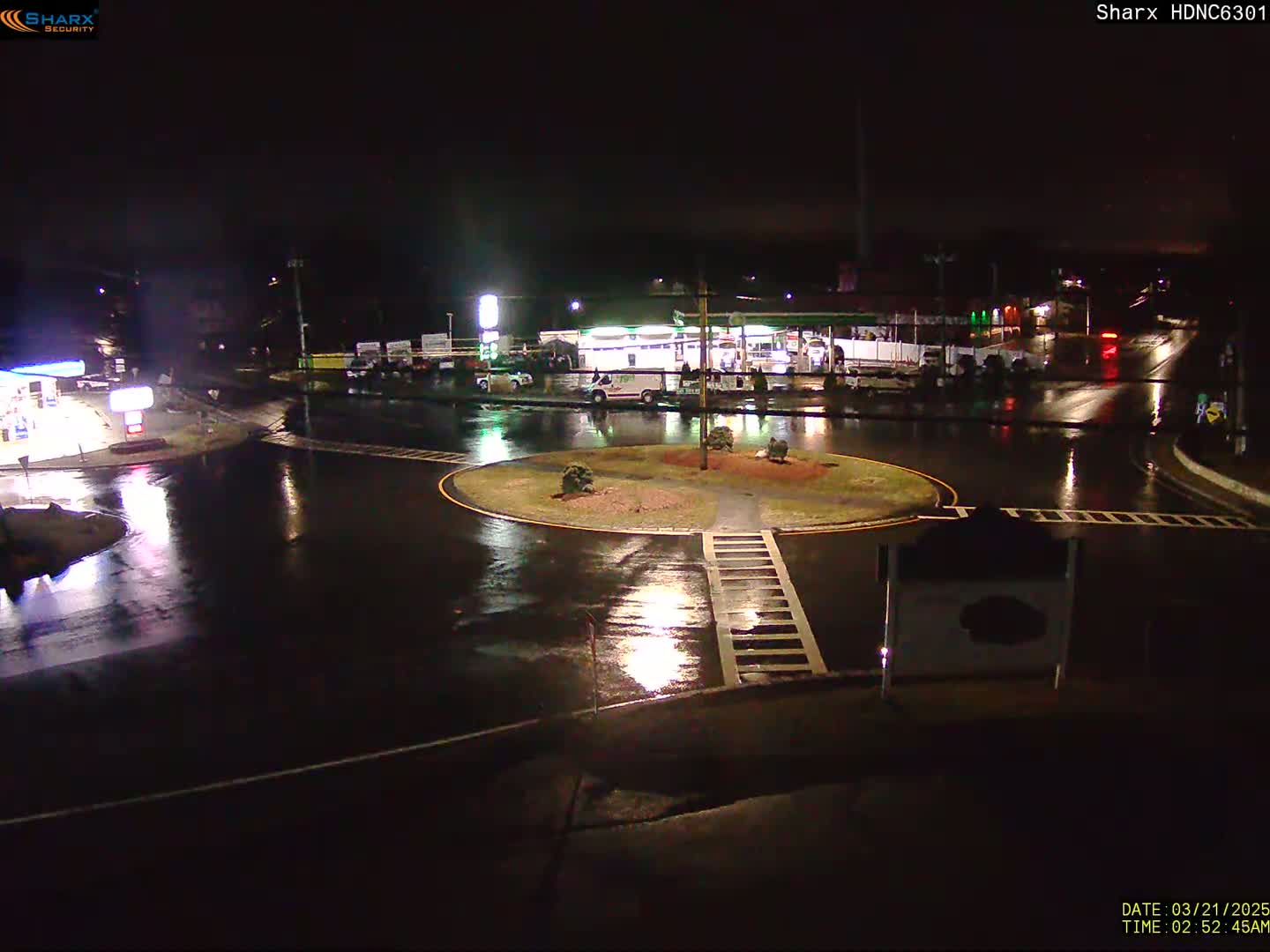 A wet, nighttime view of a roundabout intersection with a gas station and sparse traffic.