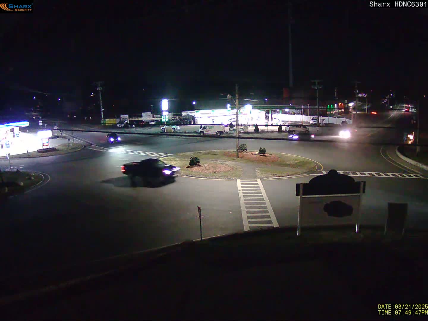 A nighttime view of a roundabout with several vehicles, including a pickup truck, and a gas station visible in the background.