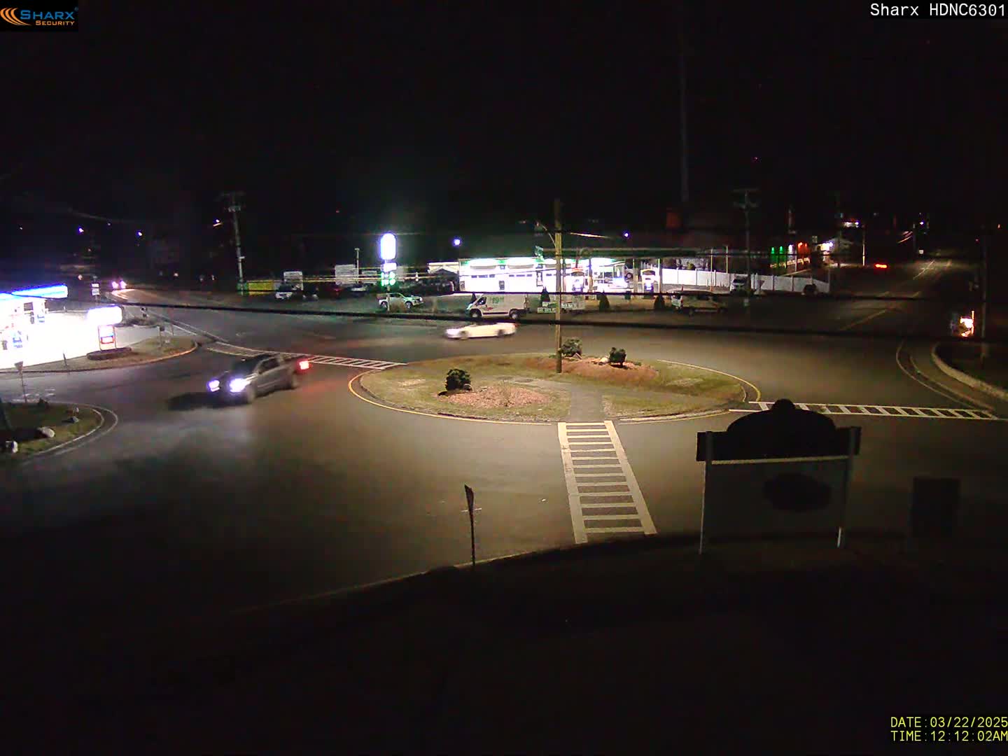 At night, a few cars navigate a roundabout near a gas station and other businesses under clear skies.