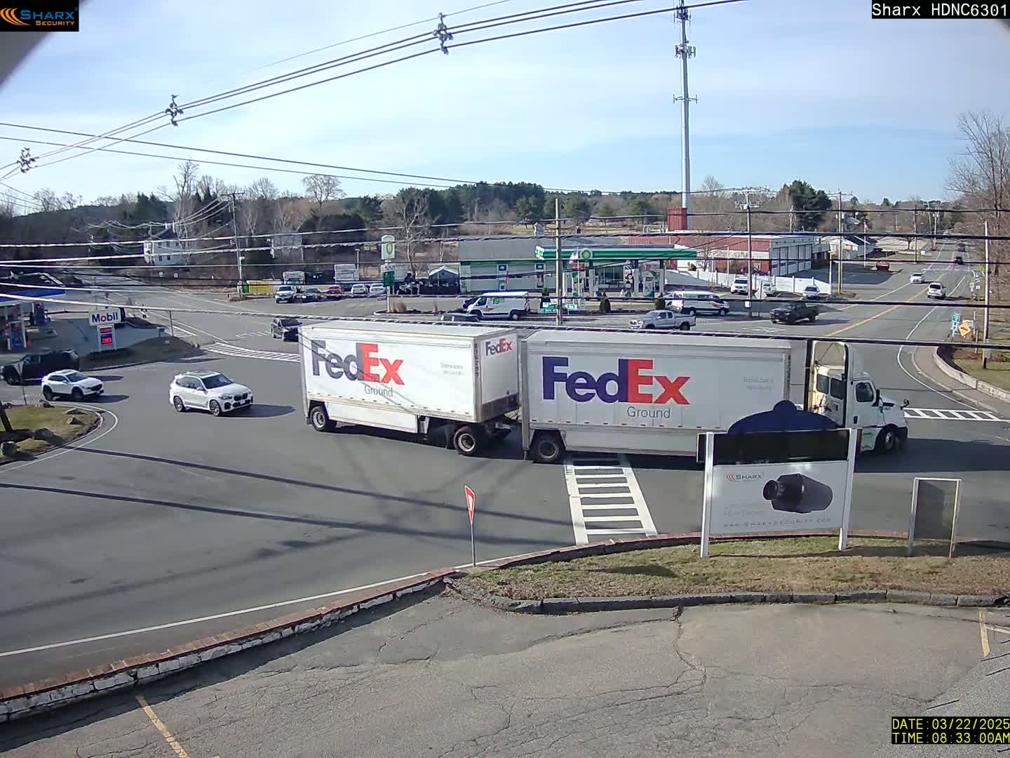 Under a sunny sky, two FedEx Ground trucks are parked at a crossroads near a gas station and other vehicles.