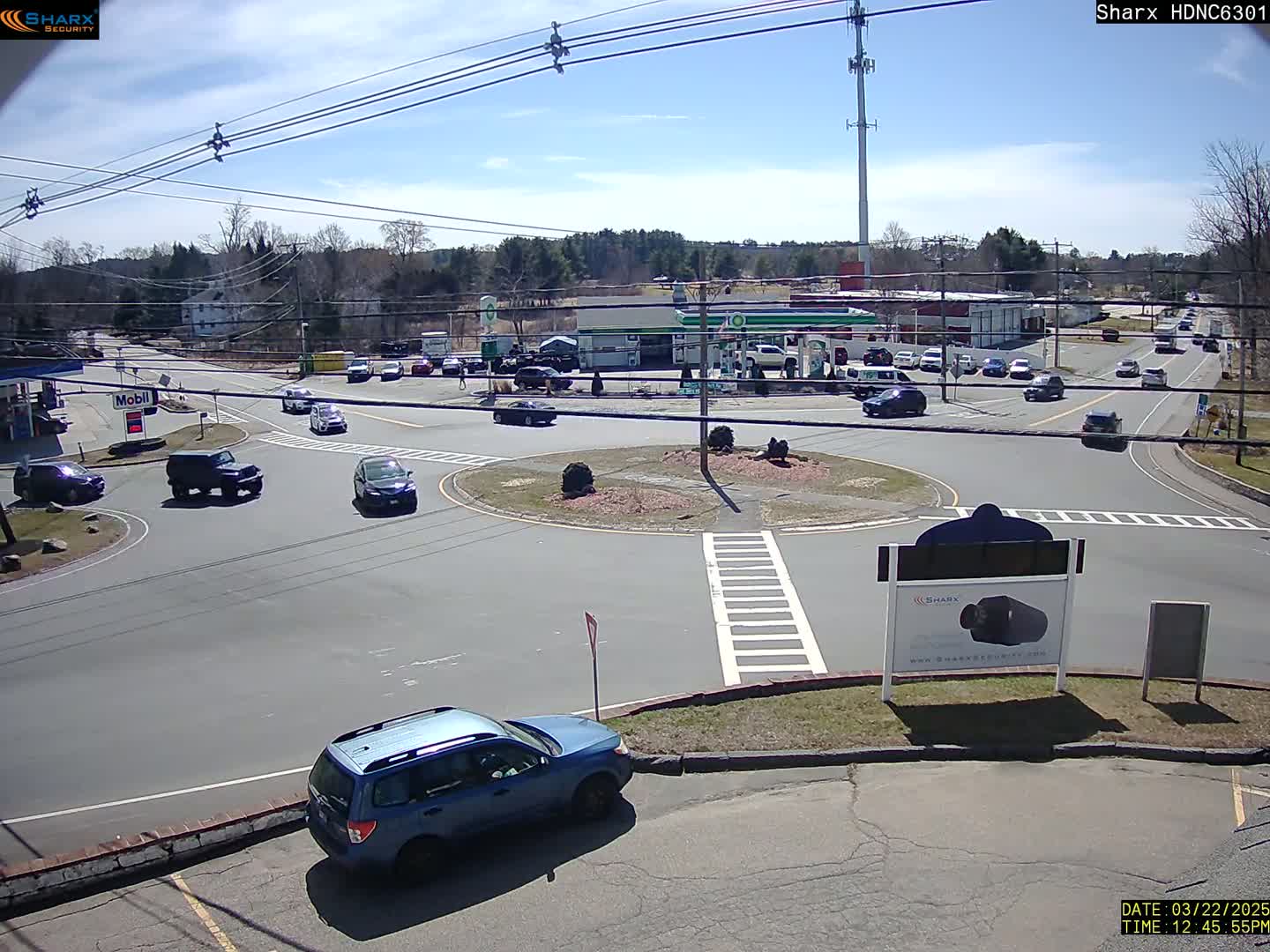 A sunny day shows a traffic circle with several cars, a gas station, and some sparse trees in the background.